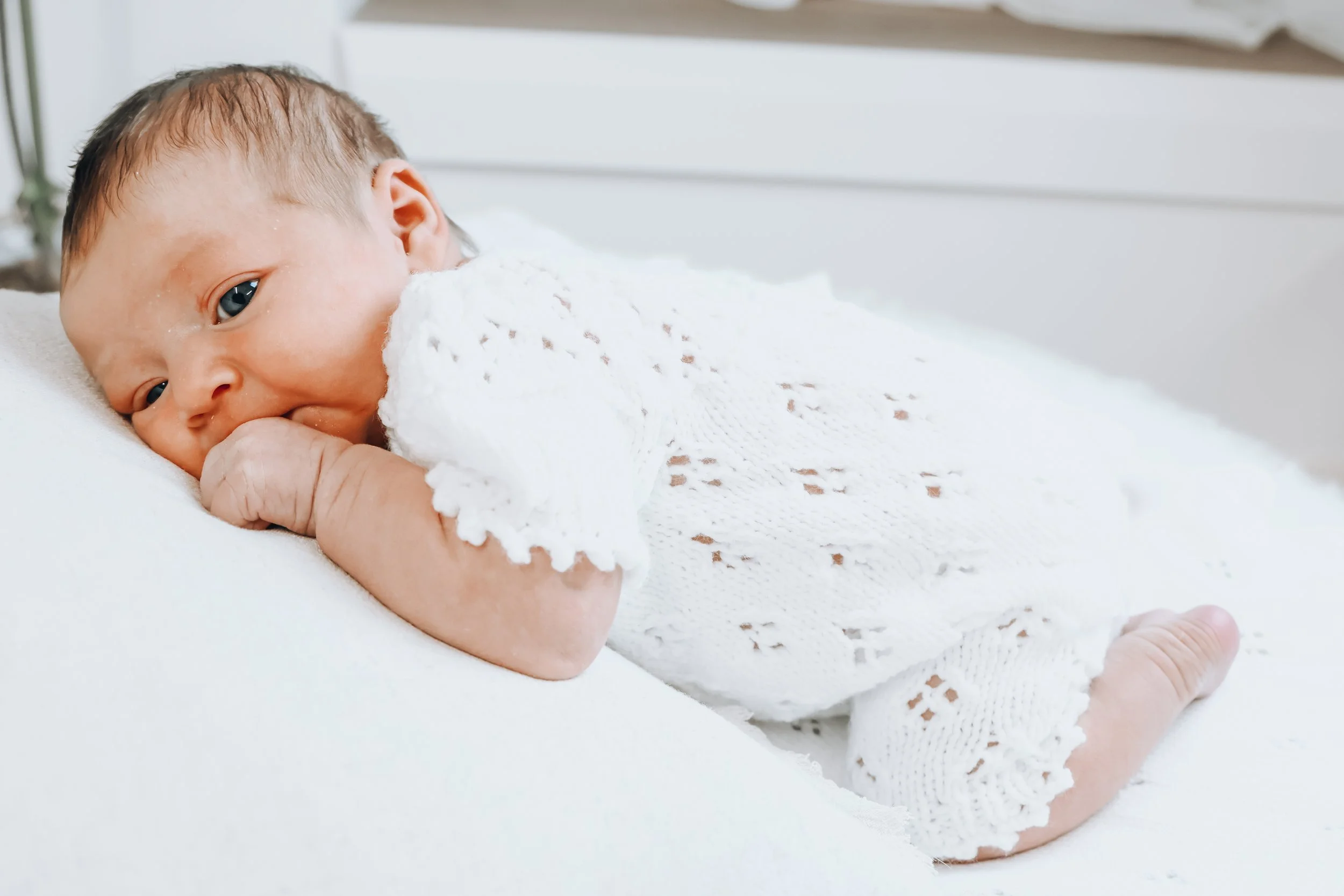 Close-up of a newborn baby lying on a white blanket, gazing at the camera with blue eyes, dressed in a white knit outfit.