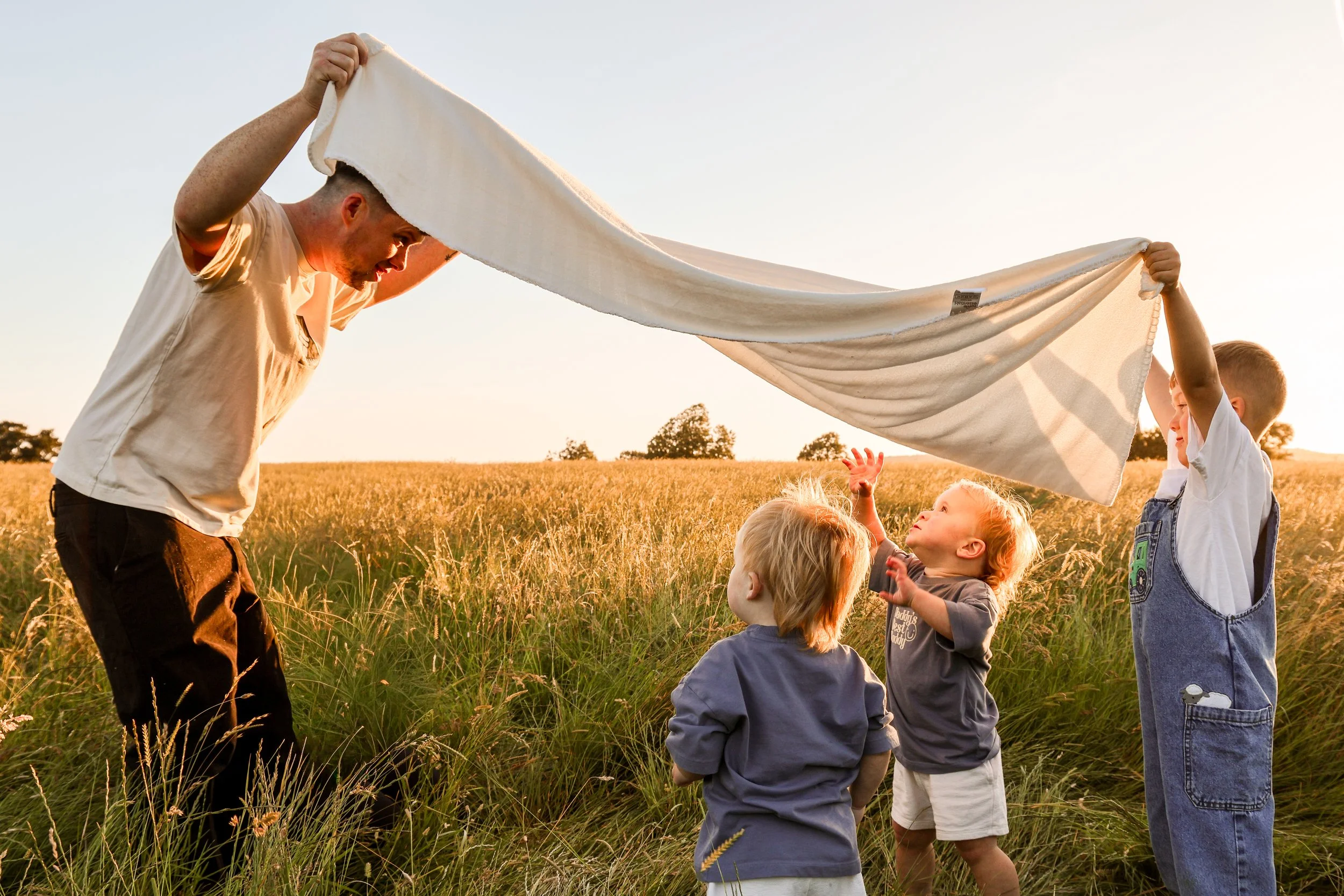 A man and five children outdoors in a field during sunset, holding a large white cloth, possibly a blanket or sheet, between them.