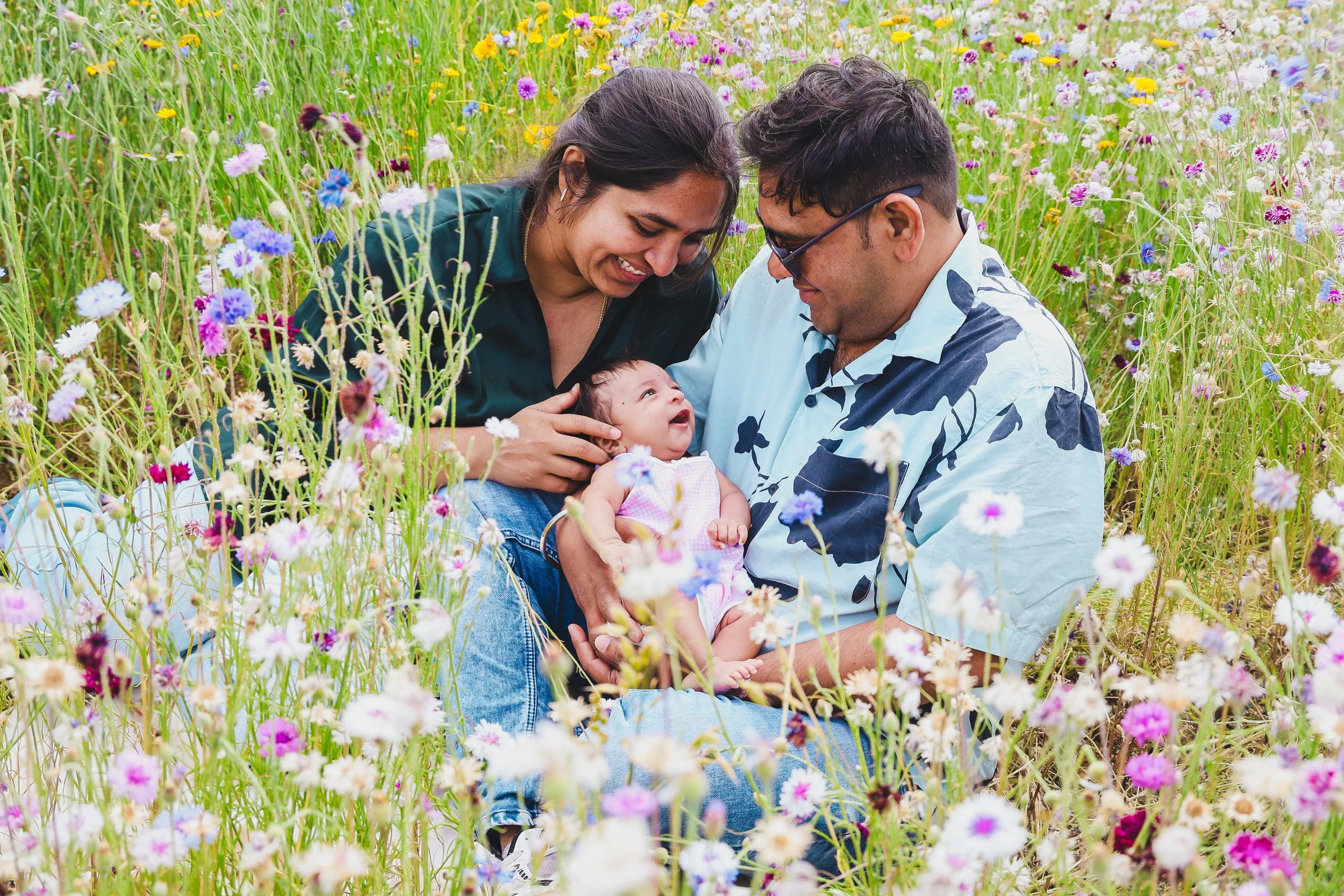 A joyful family of three, including a woman, a man, and a baby, smiles and looks at each other while sitting in a colorful wildflower meadow with blooming flowers and green grass.
