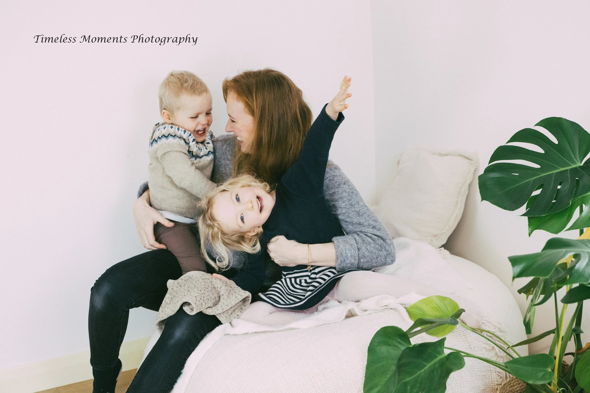 A woman with long brown hair and wearing a gray sweater is sitting on a bed with three children, playing and laughing. The children, two girls and one boy, are close to her, and they are smiling and enjoying each other's company. There is a large gre