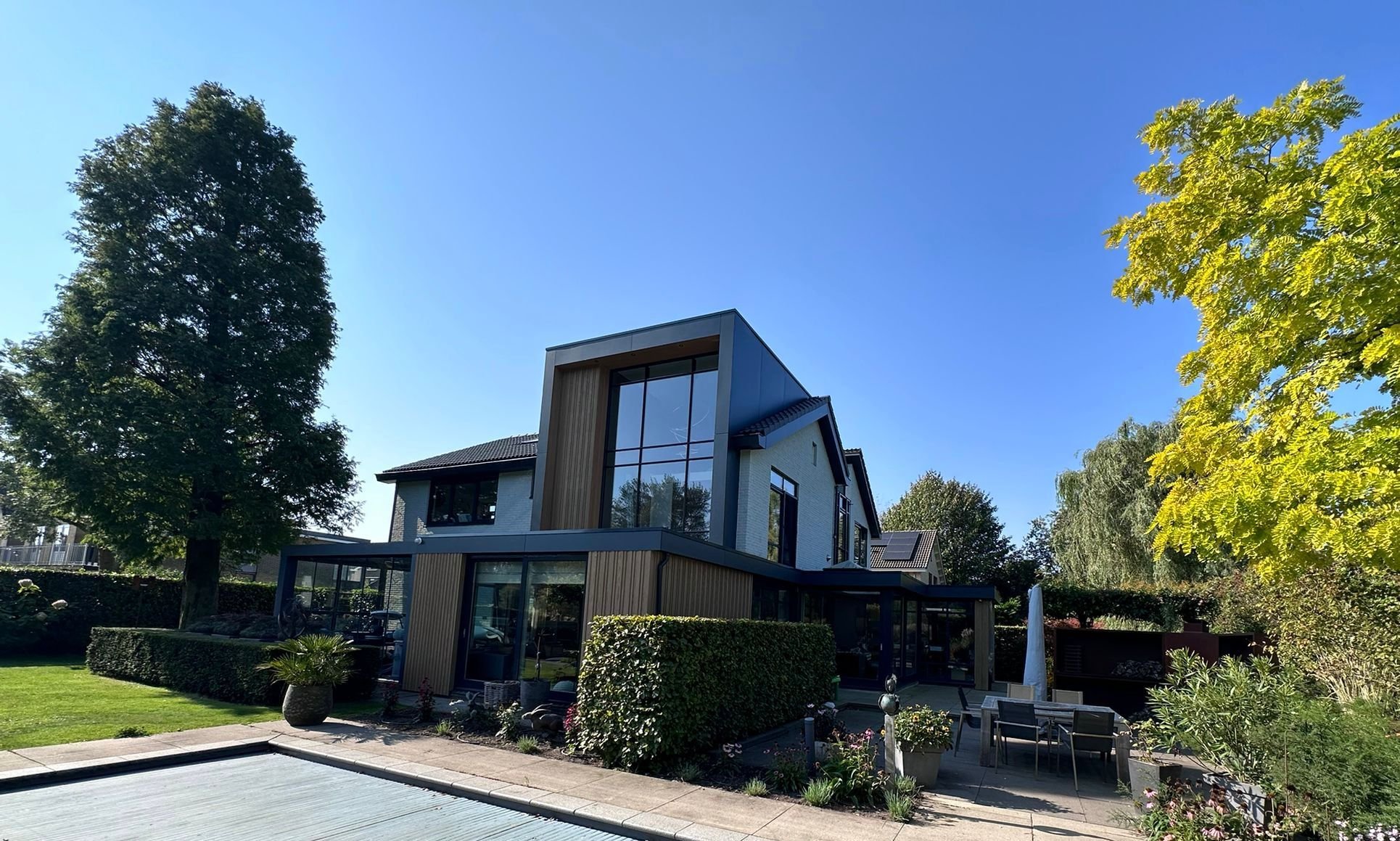 Moderne twee-verdenshuis met grote ramen, omringd door een tuin met bomen, struiken en zitgedeelte, onder een heldere blauwe lucht.