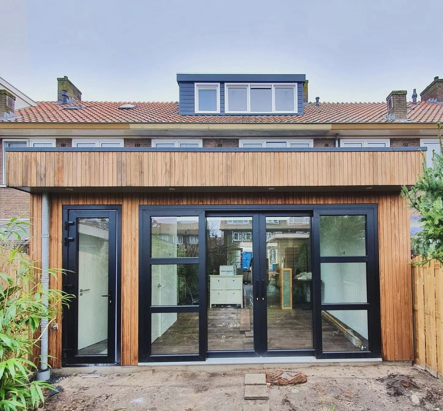 Een nieuw aangebouwde kamer of tuinkantoor met grote glazen deuren en een houten gevel, onderdeel van een rijtjeshuis, met een binnenplaats van aarde en tegels voor de ingang.