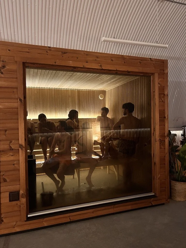 People sitting inside a wooden sauna, visible through a reflective glass window. The cozy sauna has wooden paneling and dim lighting.