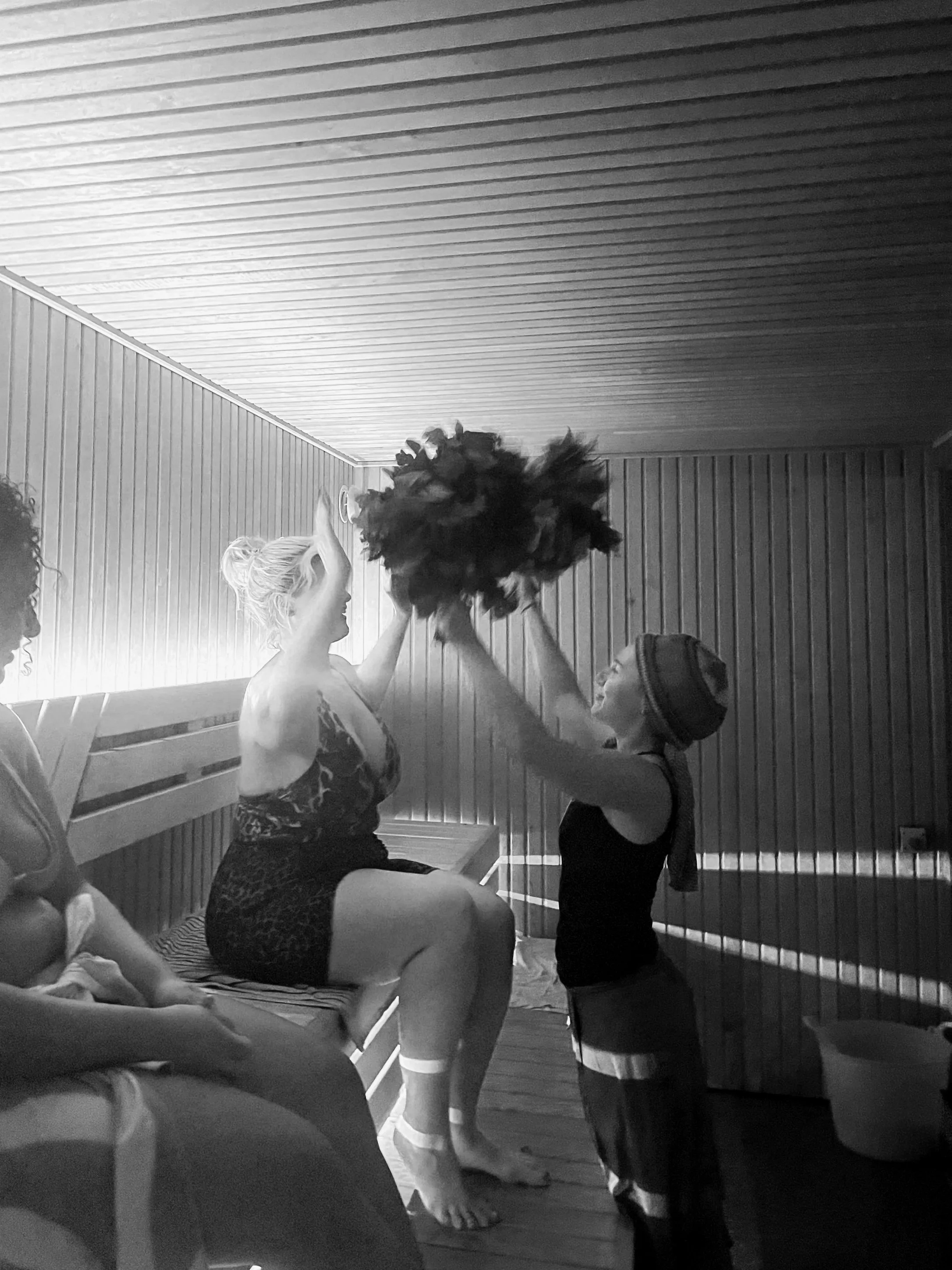 A woman in a bathing suit sits on a wooden bench in a sauna, another is holding a large bundle of leaves which she is using to pat the seated woman in a guided sauna session for women
