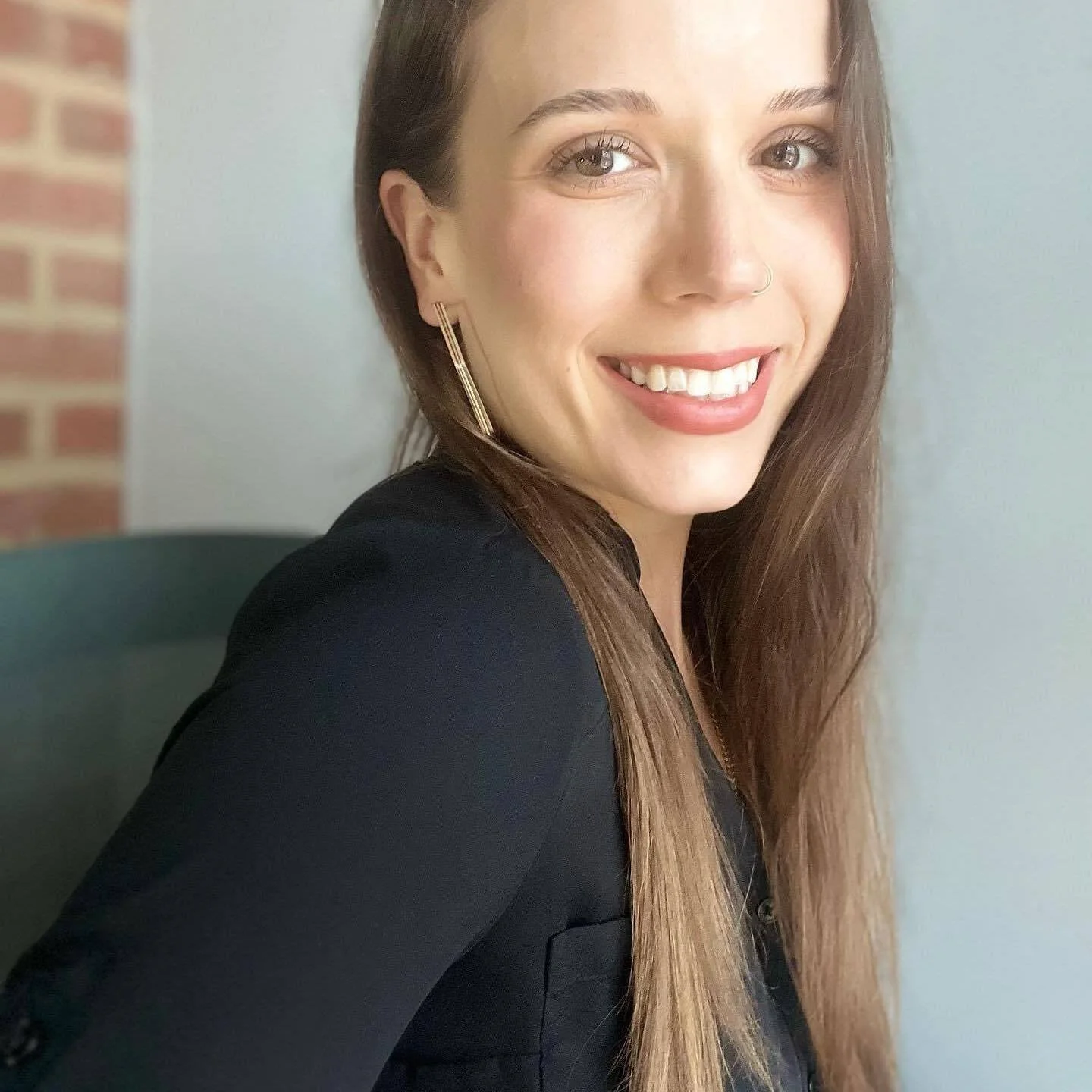 A young woman with long brown hair, light skin, and bright eyes smiling at the camera. She is wearing a black top and gold earrings, with a nose ring visible. The background includes a brick wall and neutral-colored wall.
