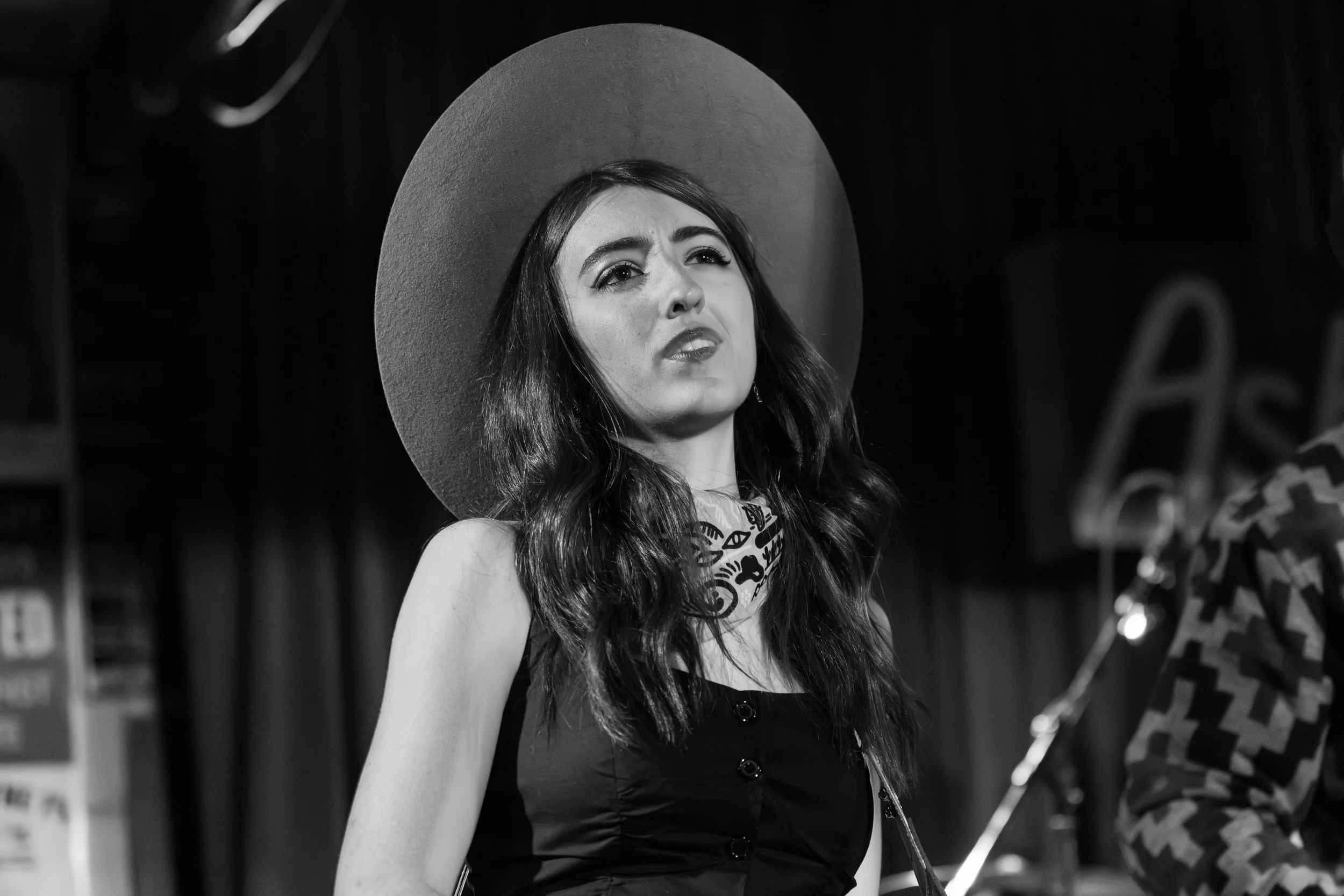 Black and white photo of Bella Rayne, a woman performing on stage, wearing a large hat and a patterned neck scarf.