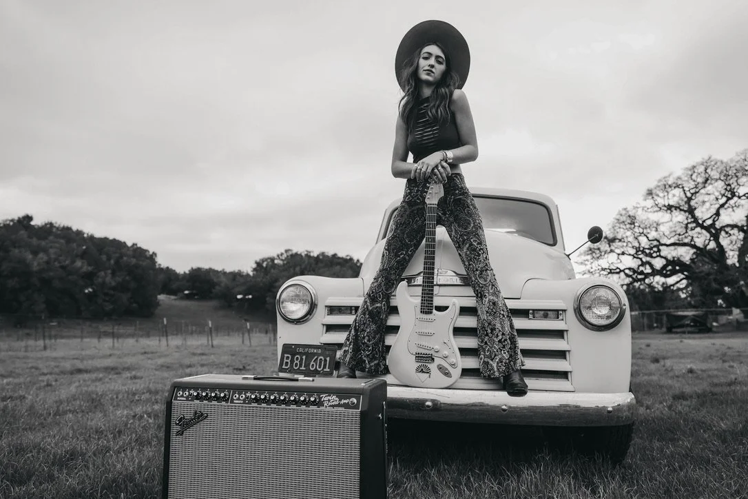 Black and white photo of Bella Rayne sitting on the hood of an old truck, holding a guitar. An amplifier is placed in front of the vehicle on grass. The scene includes trees and cloudy sky in the background.
