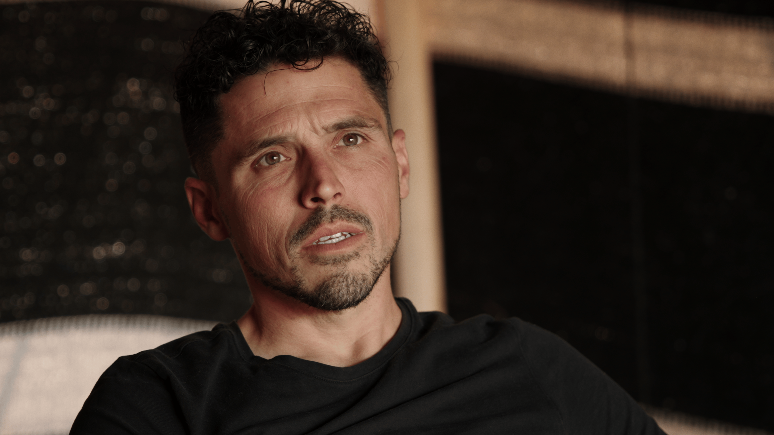 Close-up of a man with short curly black hair, light skin, and a trimmed beard, wearing a black shirt, looking confused or concerned indoors.