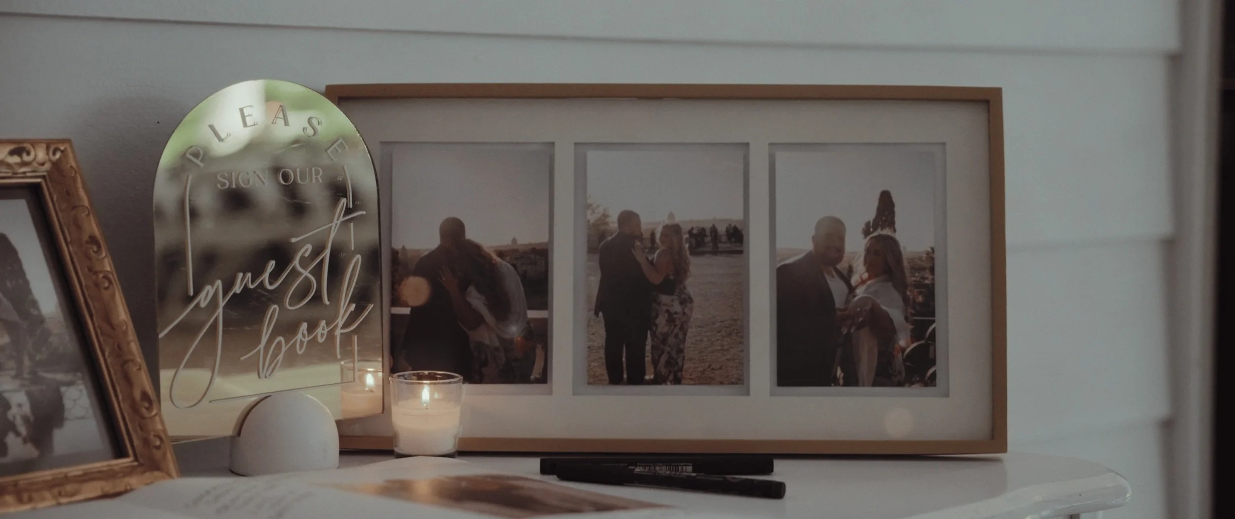 A photo collage of outdoor wedding moments behind a glass sign that reads 'Please Sign Our Guest Book' with candles and picture frames on a white table.