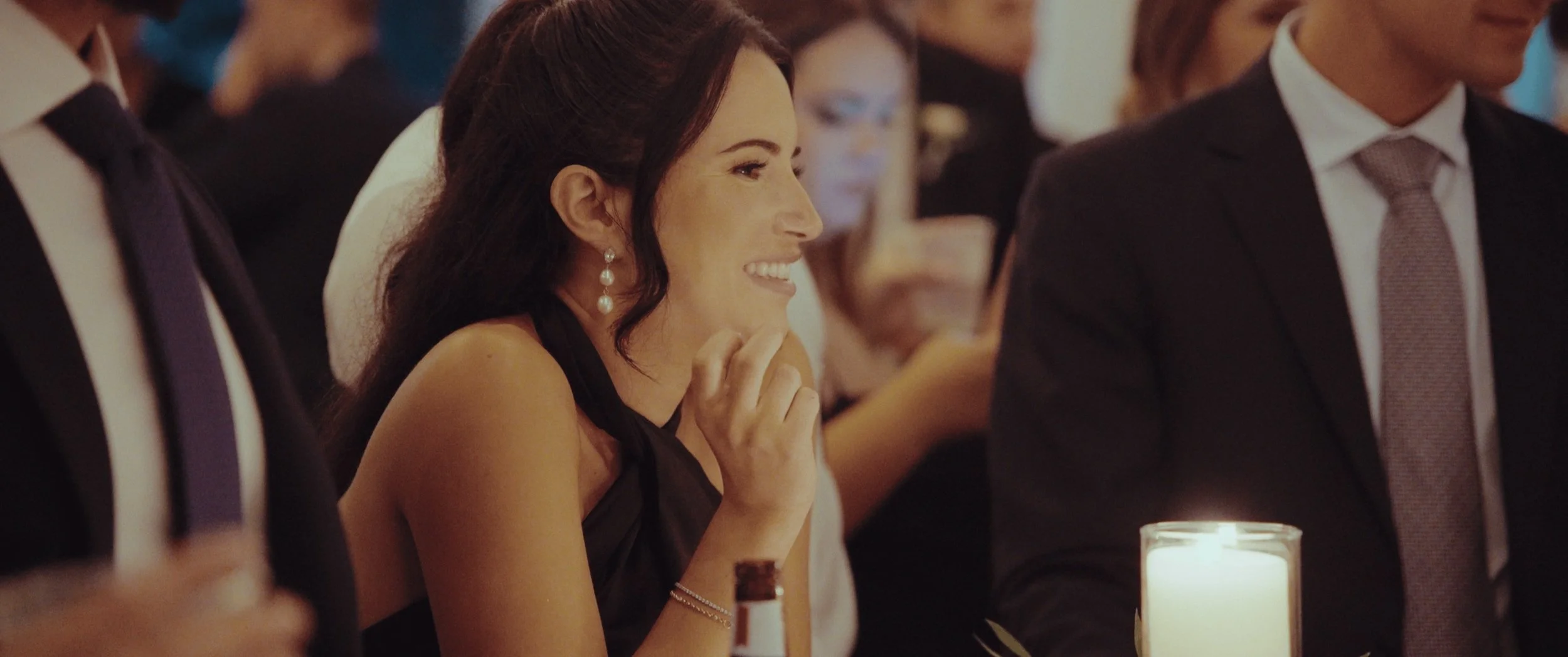 A woman smiling and laughing at a formal event or dinner, sitting next to a man in a suit, with a candle on the table in front of them.