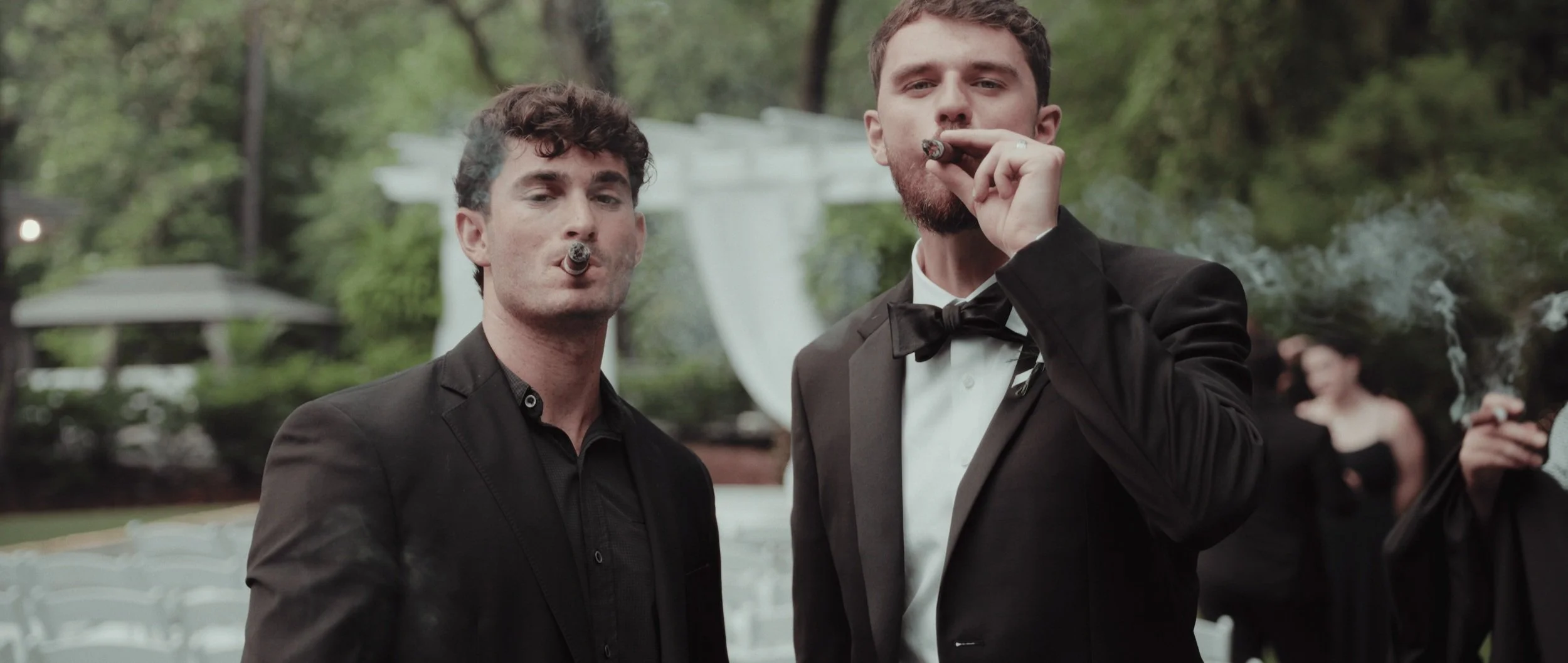 Two men in tuxedos smoking cigars at an outdoor event with greenery and white chairs in the background.