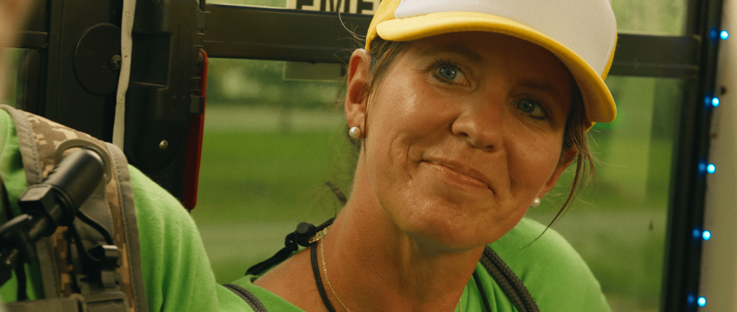 A woman with blue eyes, tan skin, and pearl earrings is smiling slightly while looking to the side. She is wearing a yellow and gray cap and a green shirt. The background is blurred, with some visible lights and greenery outside.