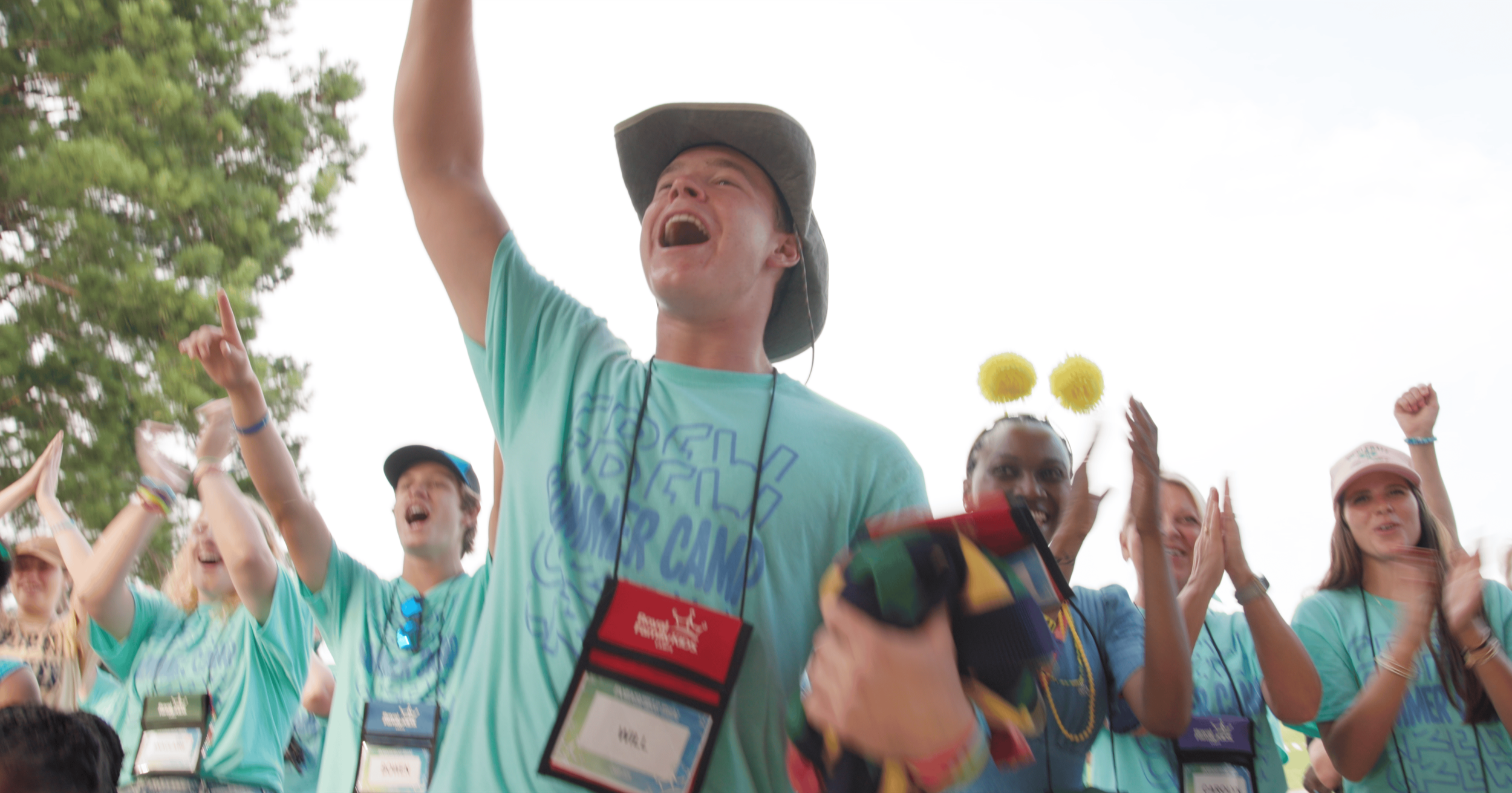 Group of happy young people celebrating outdoors, wearing conference or event badges and matching teal t-shirts, with some raising their hands and smiling, during daytime.