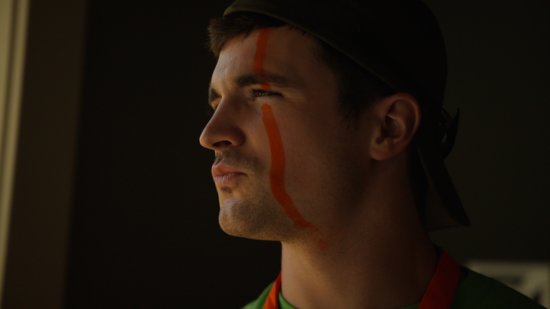 Side profile of a man with a painted red line on his face, wearing a black cap and a rainbow-colored shirt, looking out a window with sunlight illuminating his face.