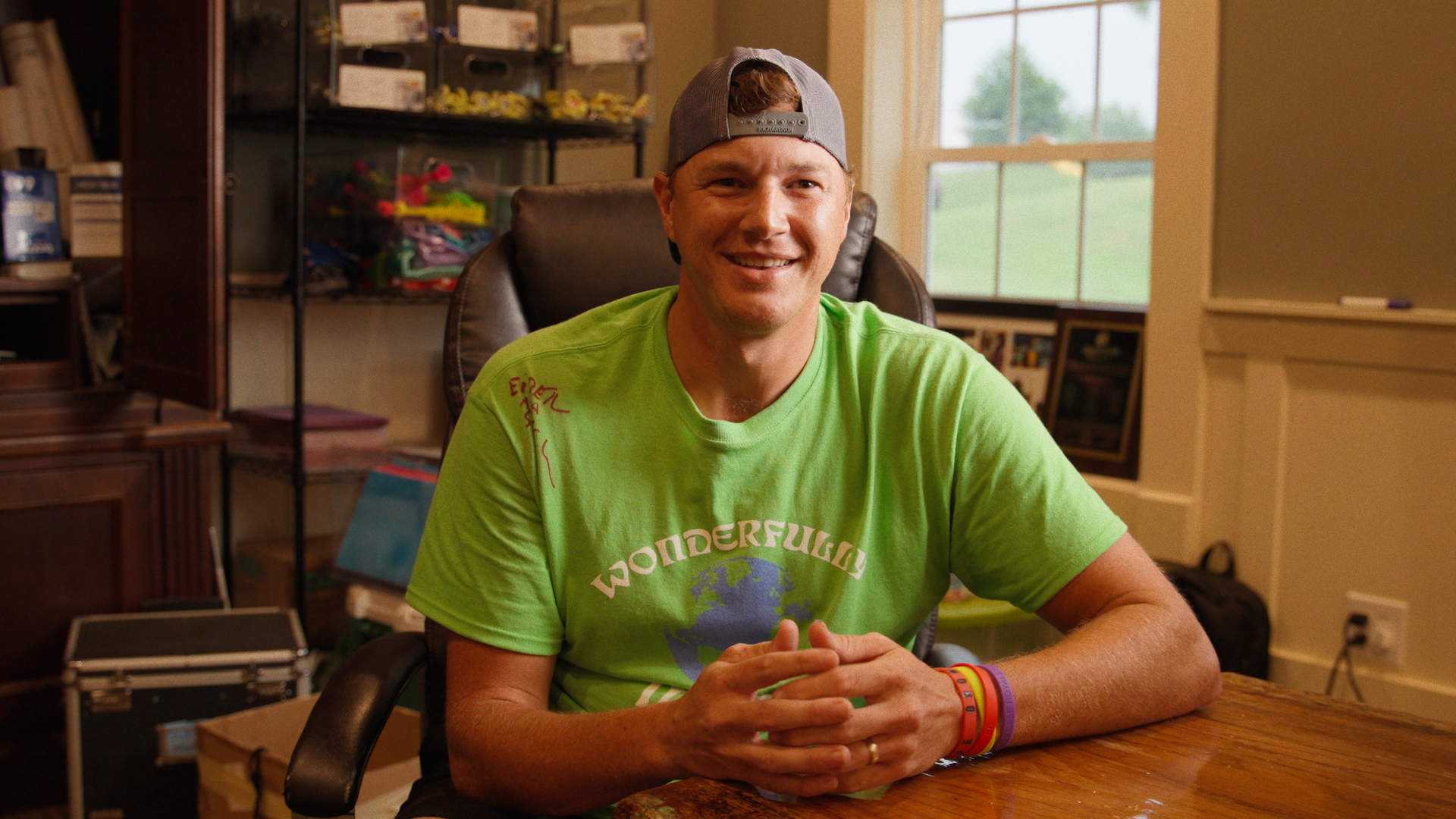 A young man with red hair wearing a grey baseball cap backwards, bright green t-shirt with a globe and the word 'Wonderful' on it, sitting at a wooden table in a room with large windows, shelves with toys and framed photos in the background, smiling 