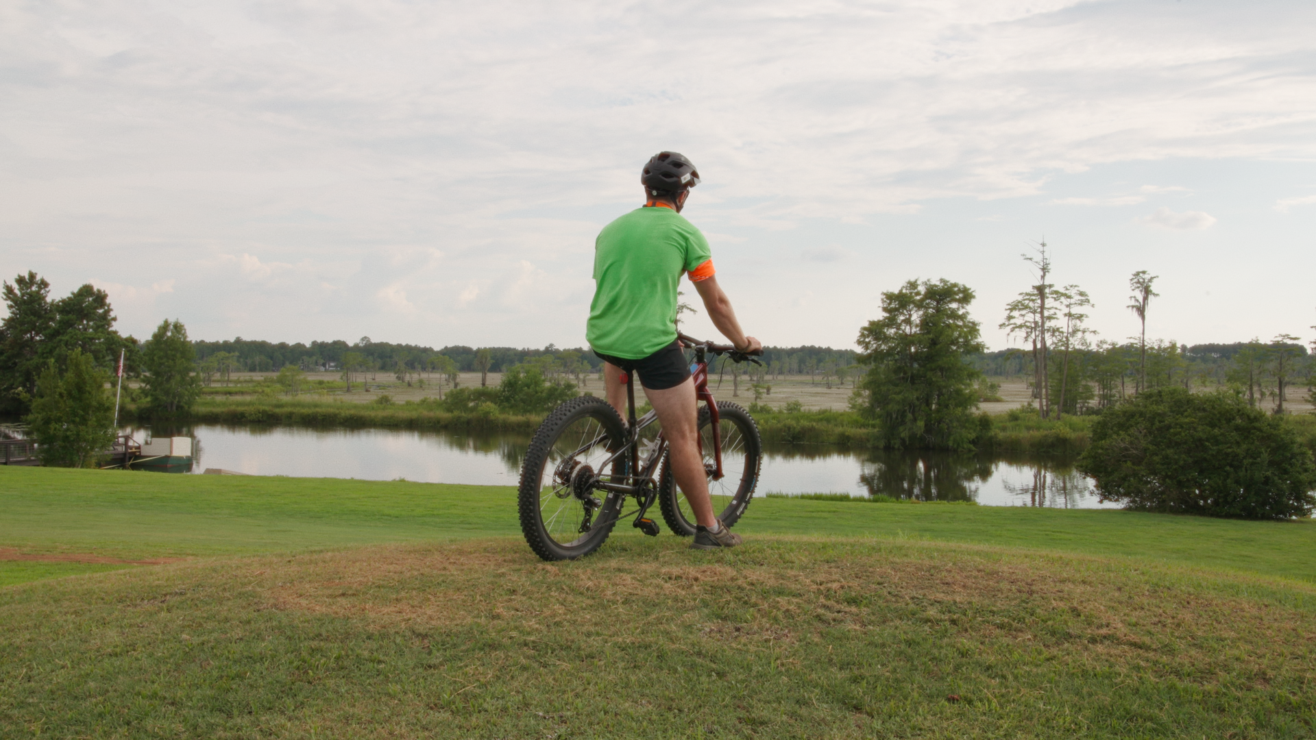 A person in a green shirt, black shorts, and helmet riding a mountain bike on a grassy area by a lake, with trees and a partly cloudy sky in the background.