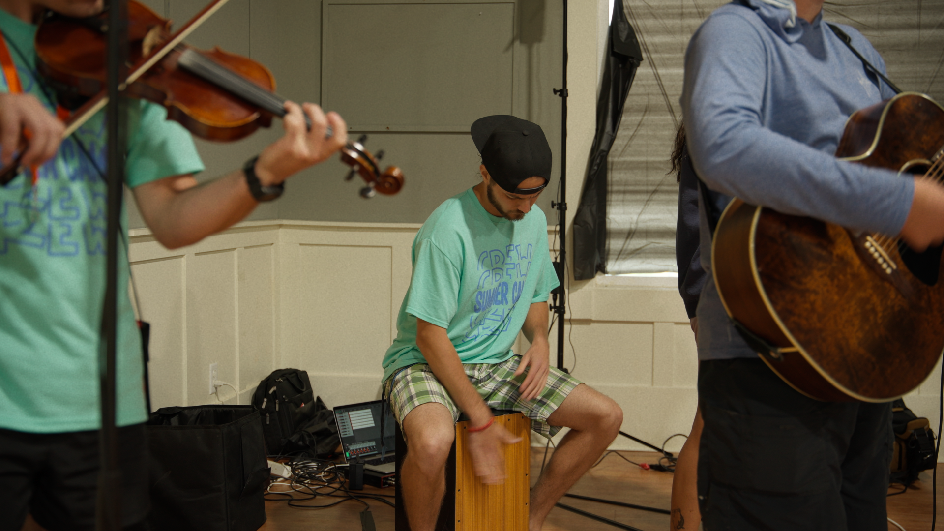 A musician sitting on a cajón drum playing with a group of other musicians playing a violin and acoustic guitar in a room with beige walls and windows.