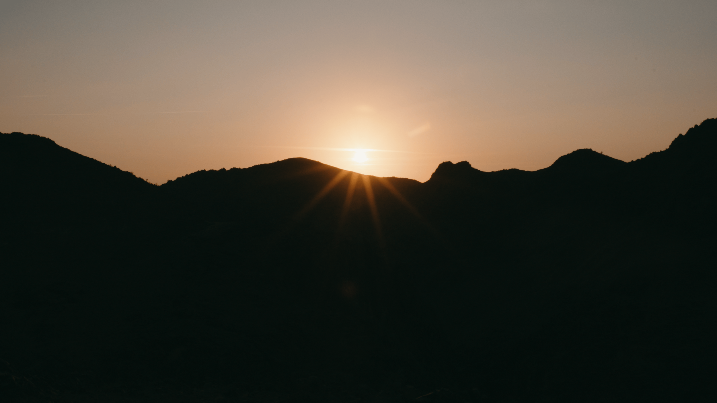 Sun setting behind mountain silhouette, with sun rays extending outward, in a peaceful landscape scene.