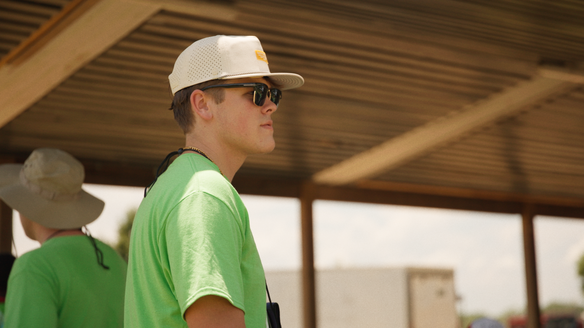 Young man in a neon green shirt, wearing sunglasses, a white cap, and a gold necklace, standing outdoors under a wooden structure.