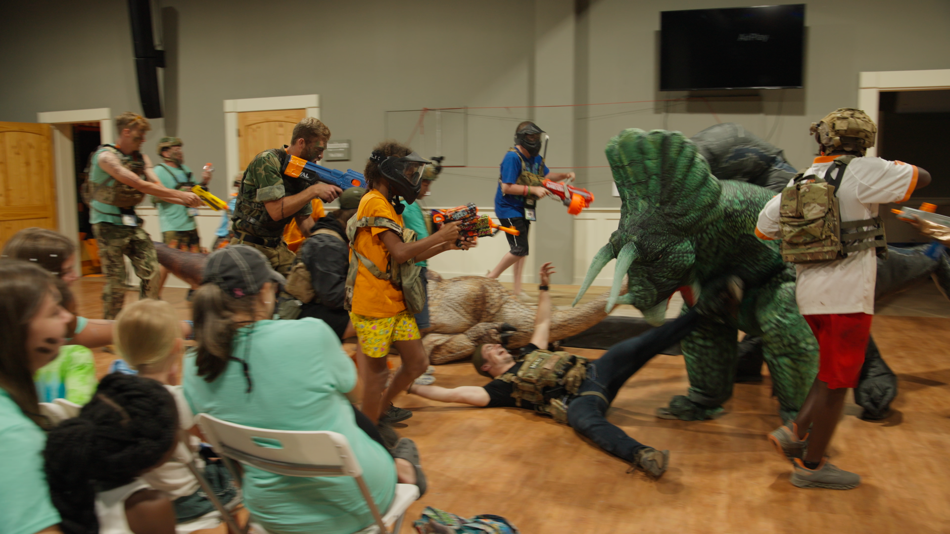 Children and adults dressed in casual and costume military gear engage in a mock battle with foam dart guns, with some in dinosaur costumes, inside a room with wooden floors and chairs.