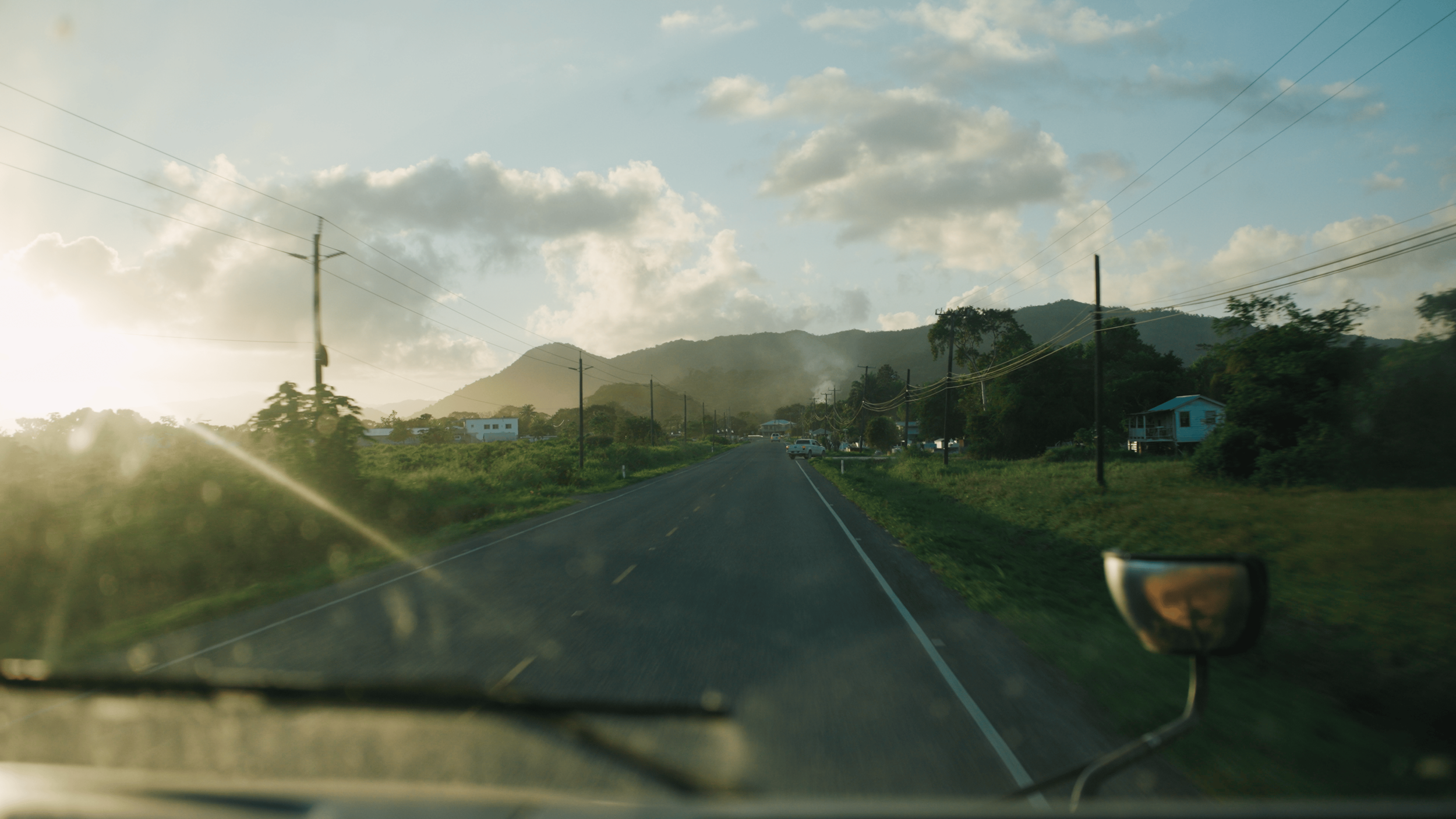 A rural road with mountains in the background, power lines on both sides, a few houses and parked cars along the roadside, and a setting sun casting a warm glow with clouds in the sky.