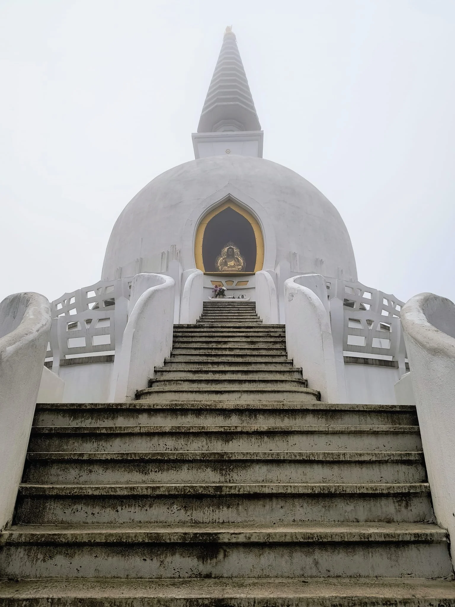 White Buddhist stupa with stairs leading up to a shrine housing a golden Buddha statue, shrouded in mist.