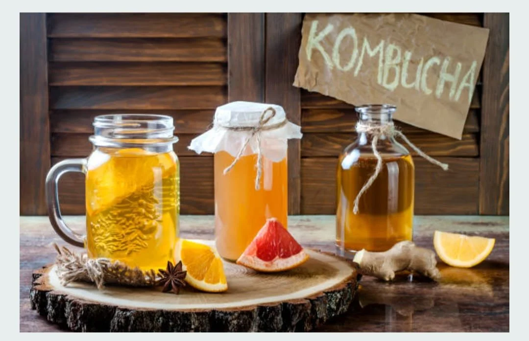 Three jars of homemade kombucha on a wooden table with citrus slices and ginger, against a wooden background with a hand-written "KOMBUCHA" sign.