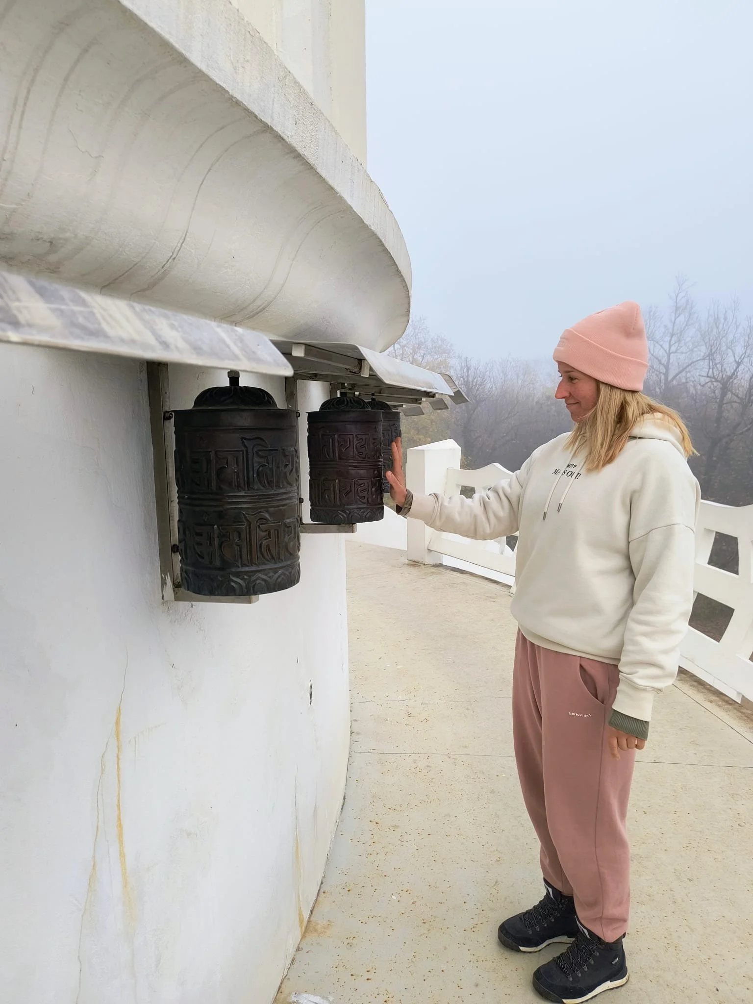 A woman wearing a pink beanie, white hoodie, pink sweatpants, and black shoes, touching two prayer wheels mounted on a white wall outside during a foggy day.