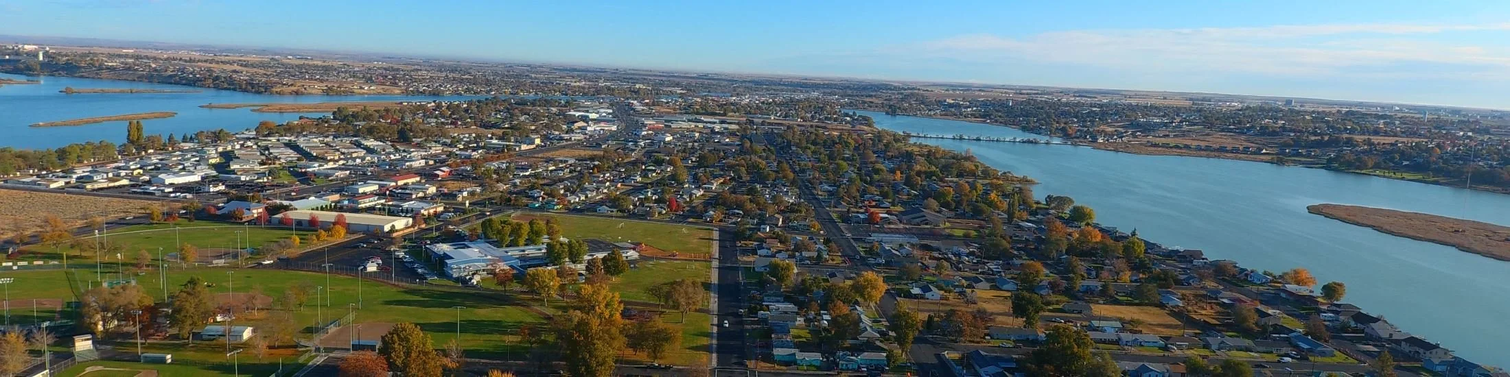 Aerial view of a suburban area with a river and green spaces, surrounded by residential houses and some commercial buildings under a clear blue sky.