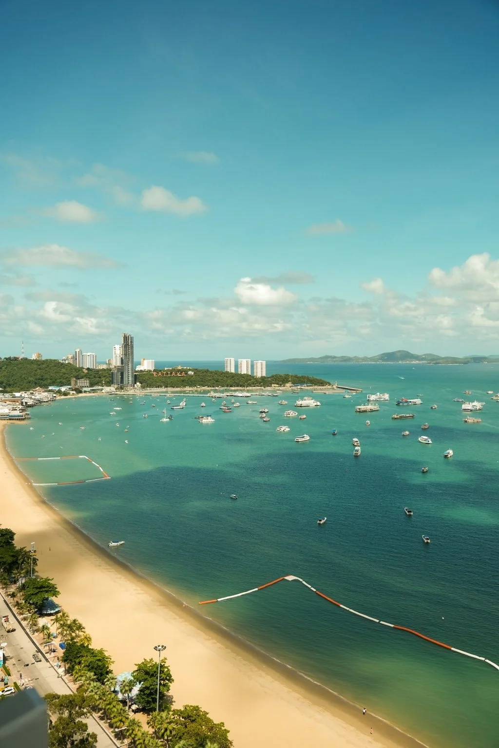 erial view of Pattaya Bay with sandy beachfront, turquoise water and boats anchored offshore under a clear blue sky.