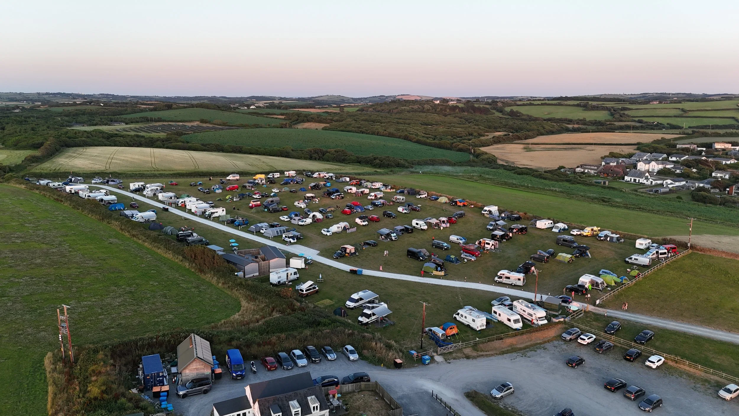 A large parking area filled with cars, RVs, and tents, surrounded by green fields, rolling hills, and a few houses in the distance during sunset.