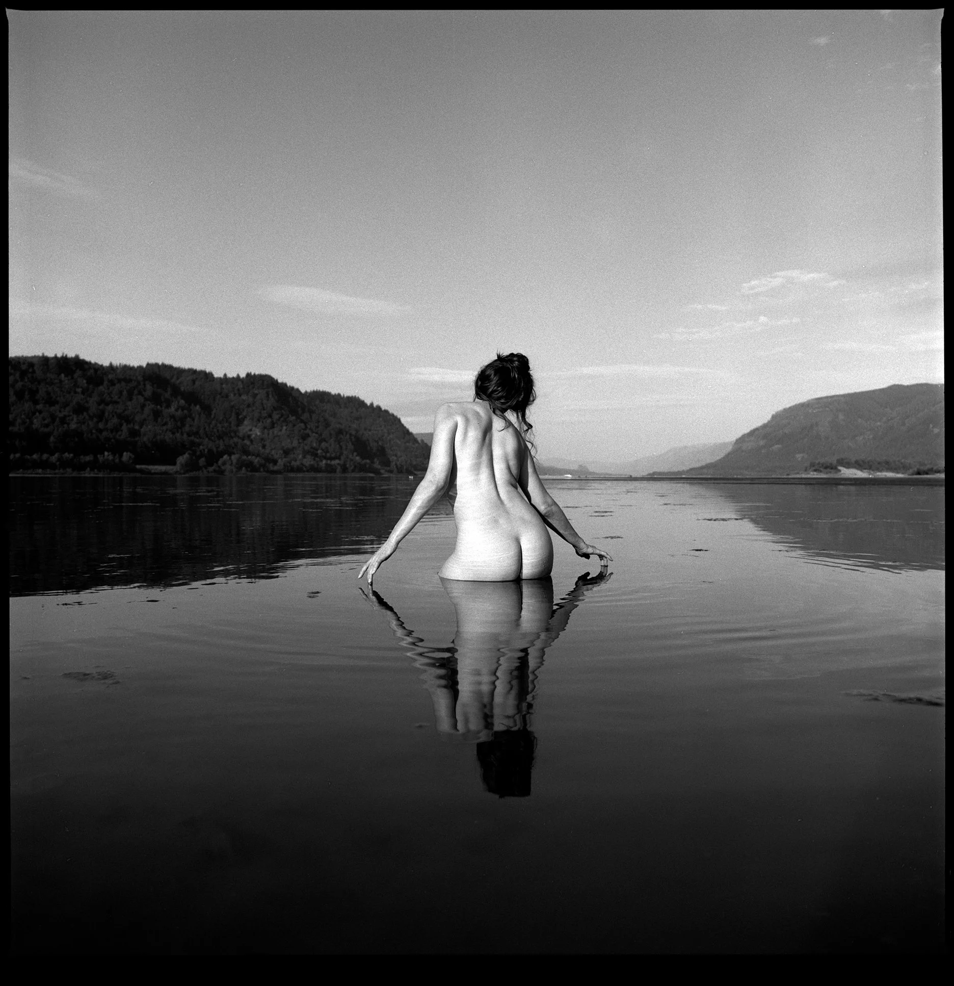 Nude woman crouching in the water with a reflection at Rooster Rock state park in Oregon on black and white film.