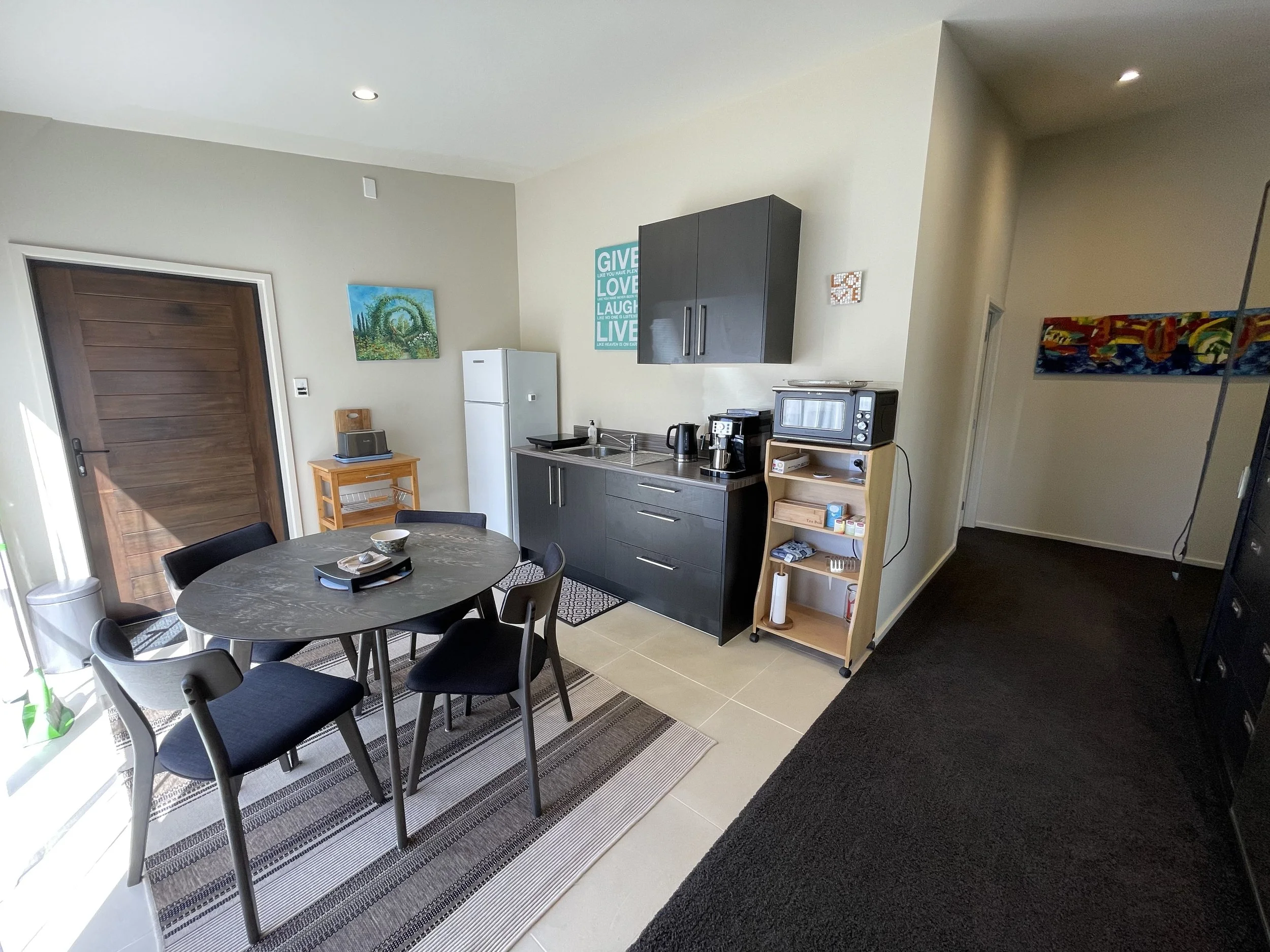 Small kitchen and dining area with a round table and four chairs, a kitchenette with a sink, microwave, and fridge, and minimalist decor with paintings on the wall. Bluewater Sanctuary Tutukaka