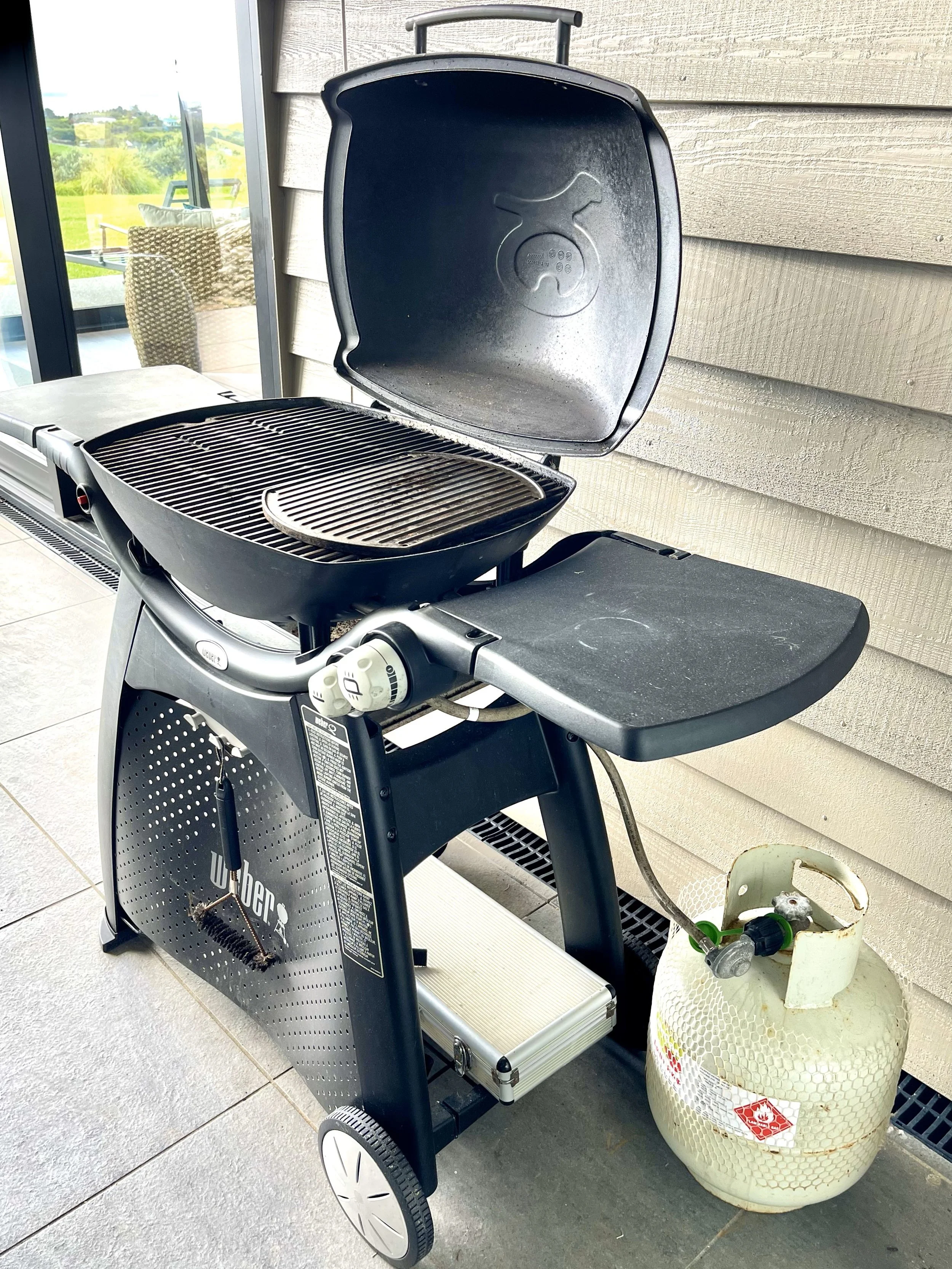 Outdoor gas grill with lid open, connected to a propane tank, sitting on a patio near a sliding glass door. Bluewater Sanctuary Tutukaka