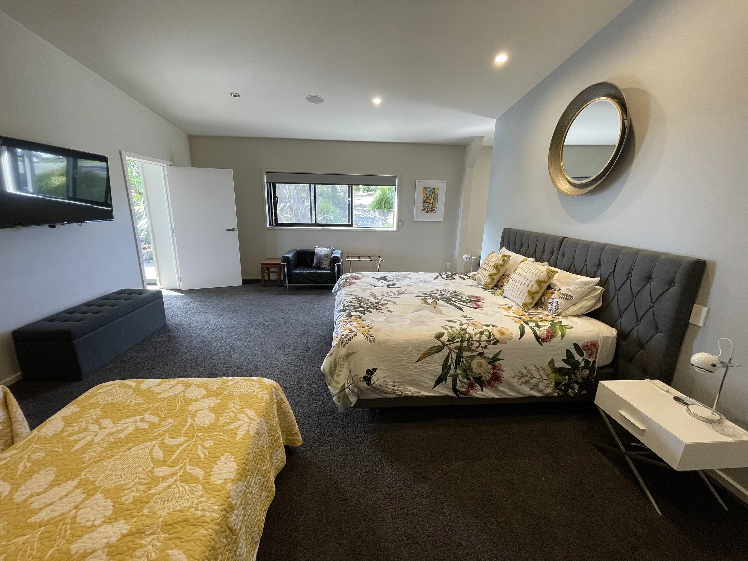 Modern bedroom with a large, floral-patterned bed, dark gray upholstered bench, flat-screen TV on the wall, and a round mirror. There is also a small armchair by the window and a yellow-covered bed in the foreground. The room has natural light, neutral walls, and minimal decor.