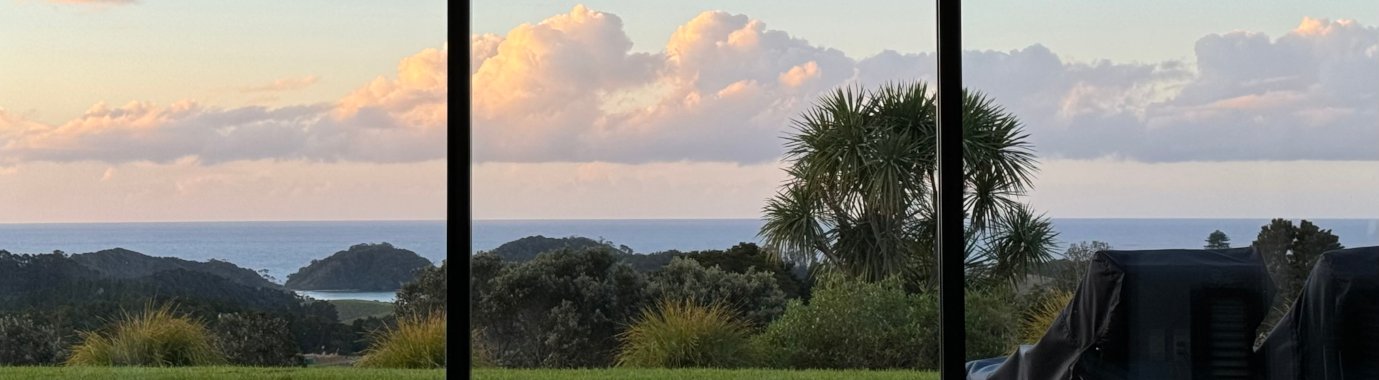 Scenic view through large windows showing a grassy area with trees, overlooking a body of water and distant hills, under a sky with fluffy clouds. Bluewater Sanctuary Tutukaka