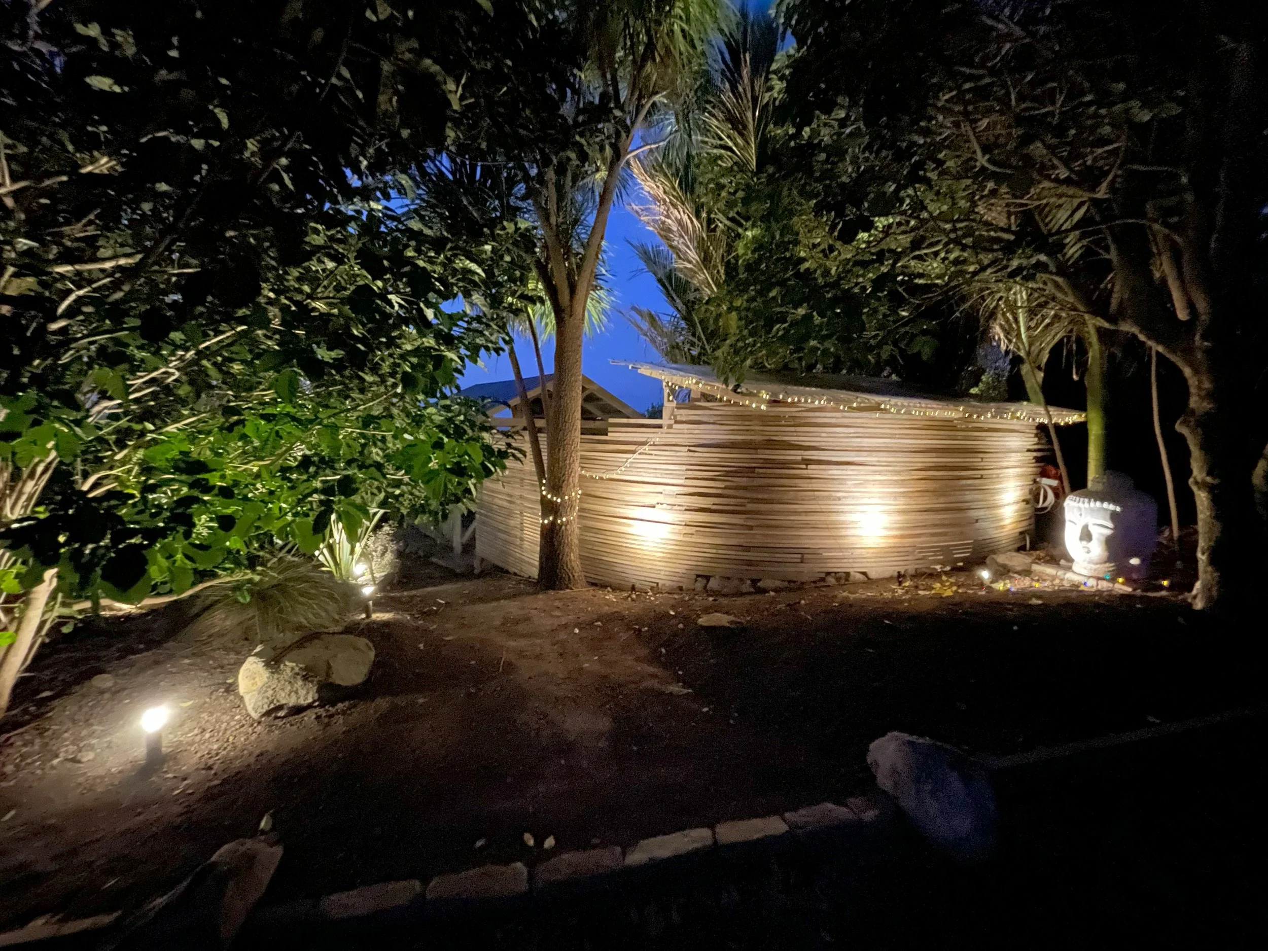 Illuminated wooden structure surrounded by trees at night, with decorative lights, a stone path, and a large sculpted face near the entrance. Bluewater Sanctuary Tutukaka
