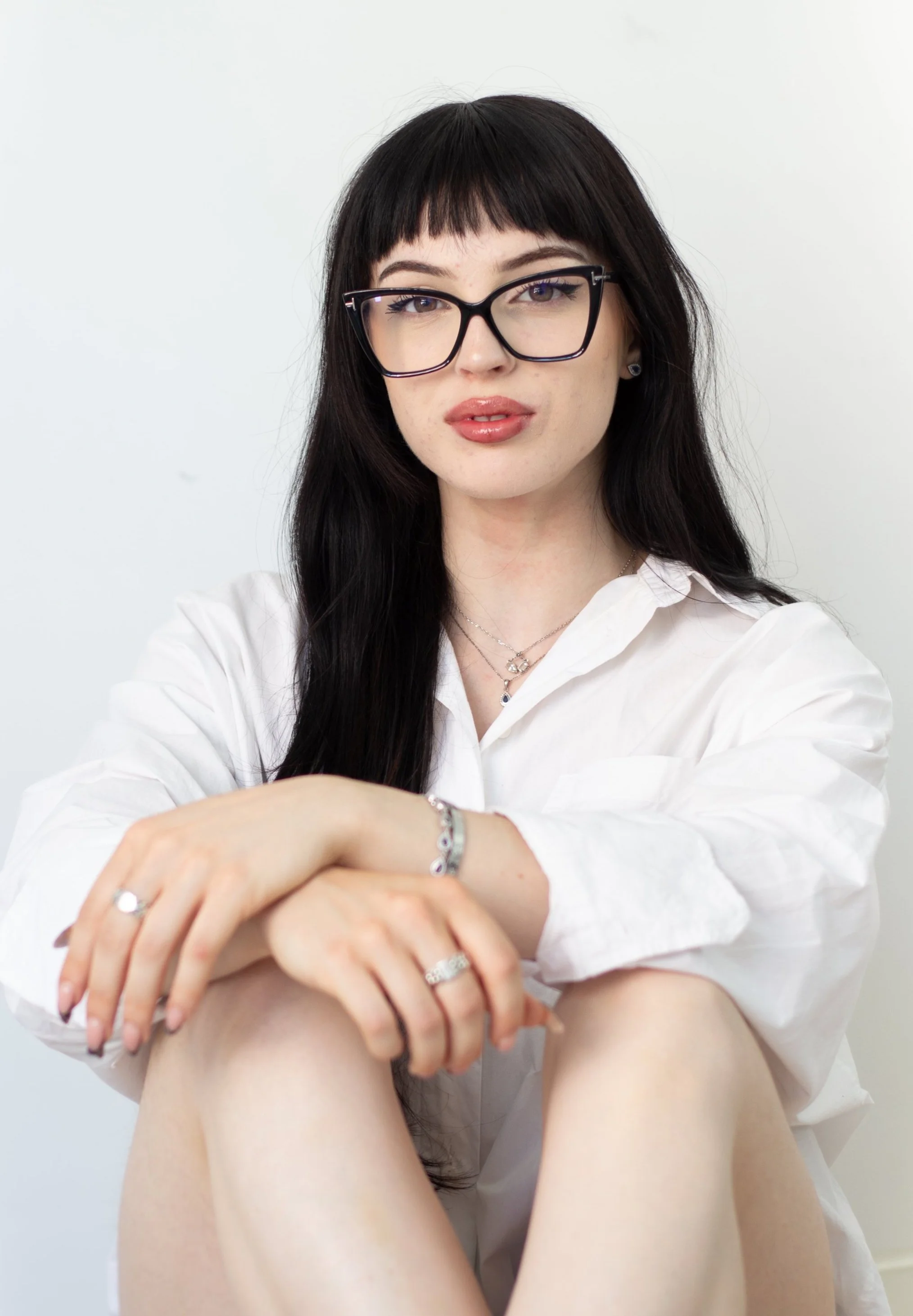 A woman with long dark hair, glasses, and light makeup, wearing a white shirt, sitting with her arms resting on her knees against a plain white background.