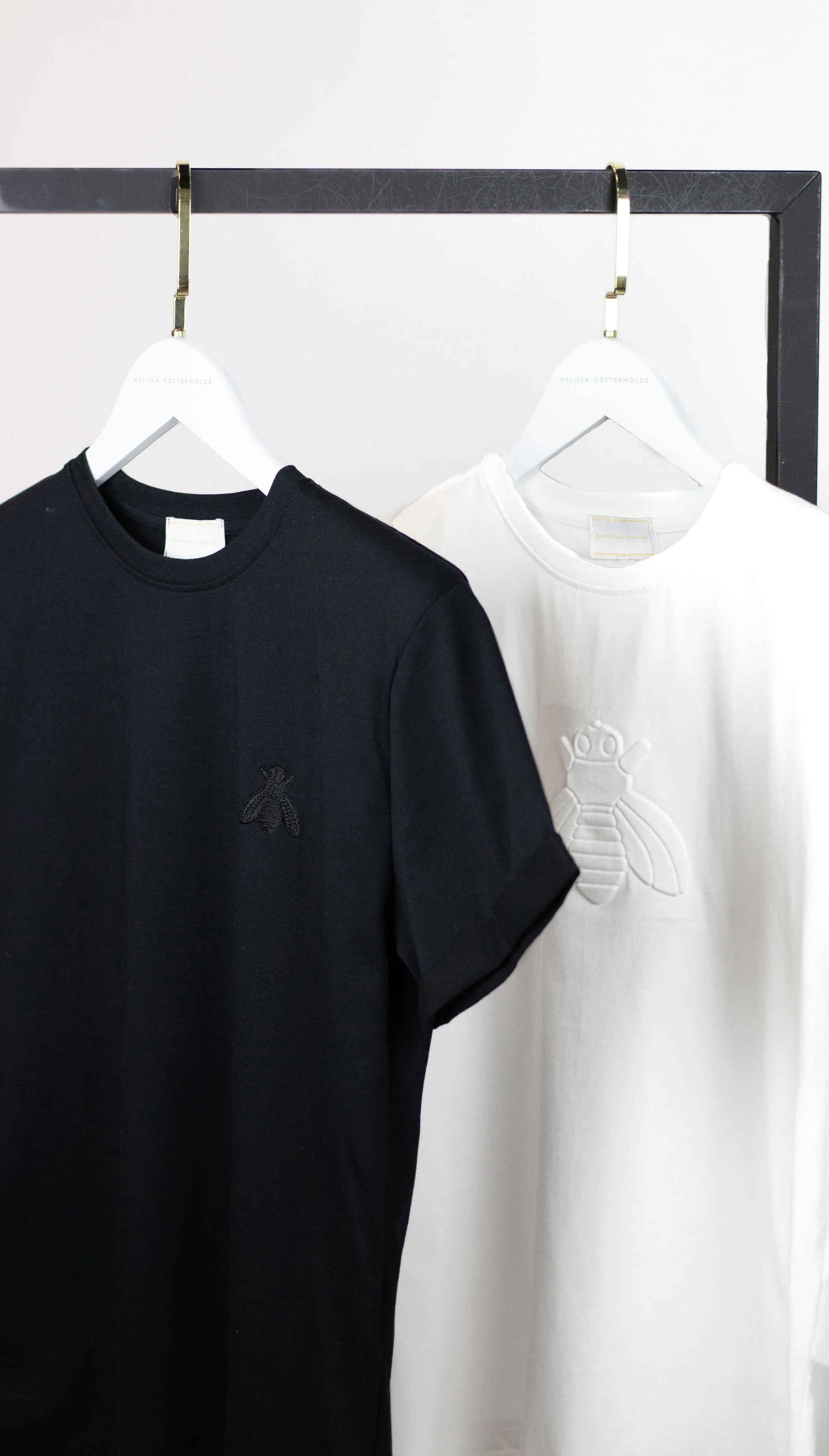 Black and white t-shirts hanging on white hangers on a black metal clothing rack. The black t-shirt has a small bee embroidered on the chest, and the white t-shirt has an embossed bee design.