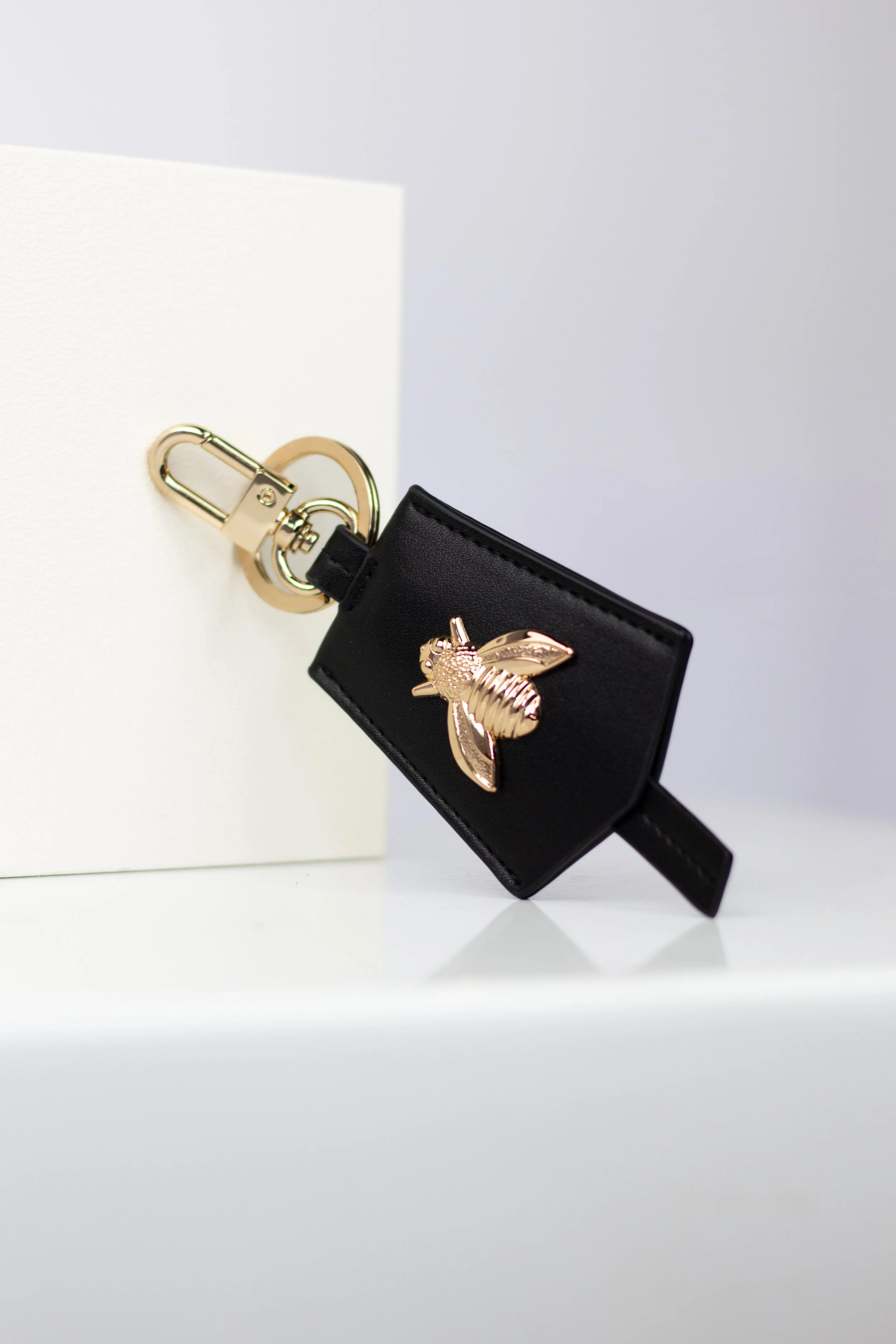 A black leather keychain holder with a gold bee emblem attached to a gold clip, hanging from a white rectangular display block against a plain background.