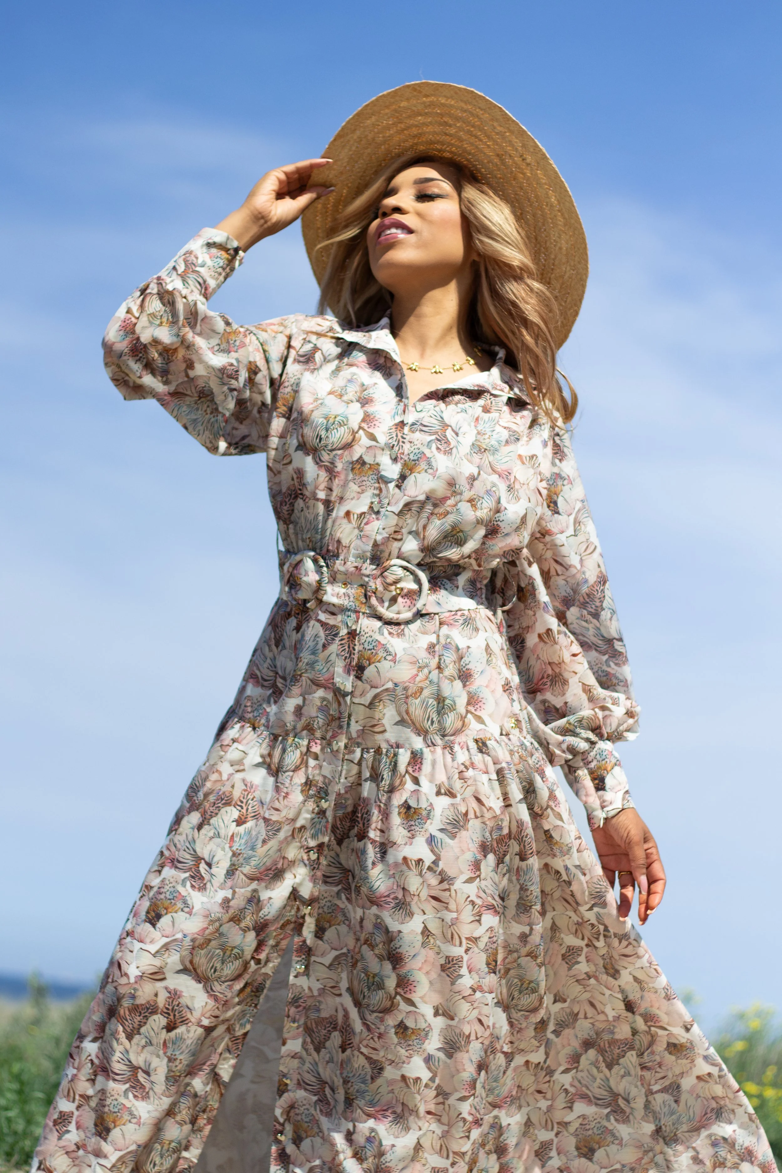 A woman wearing a floral long dress and wide-brimmed hat, standing outdoors against a blue sky.