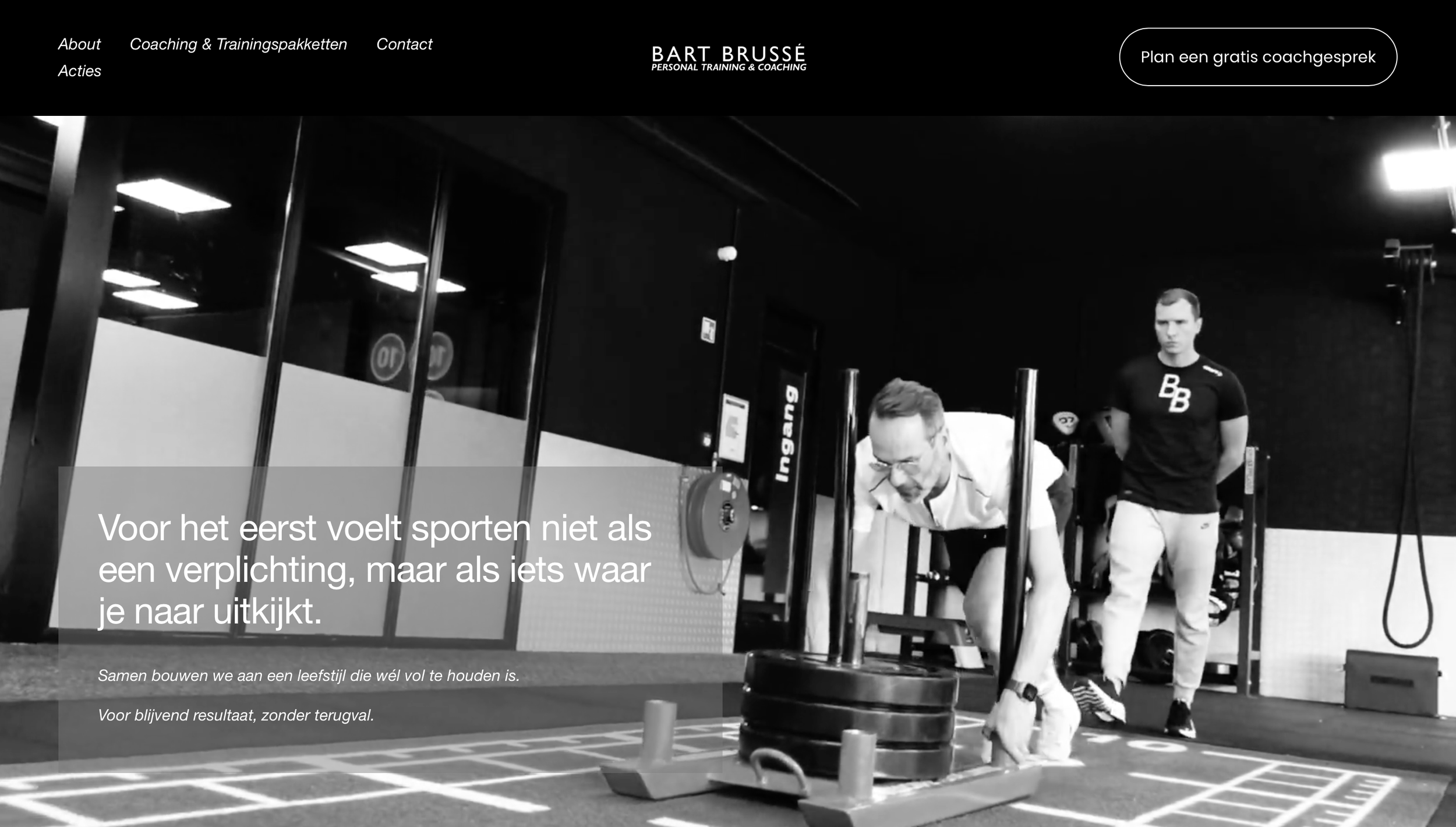 A man working out in a gym pushing a sled with weights, with a coach standing behind him watching. The scene is in black and white.