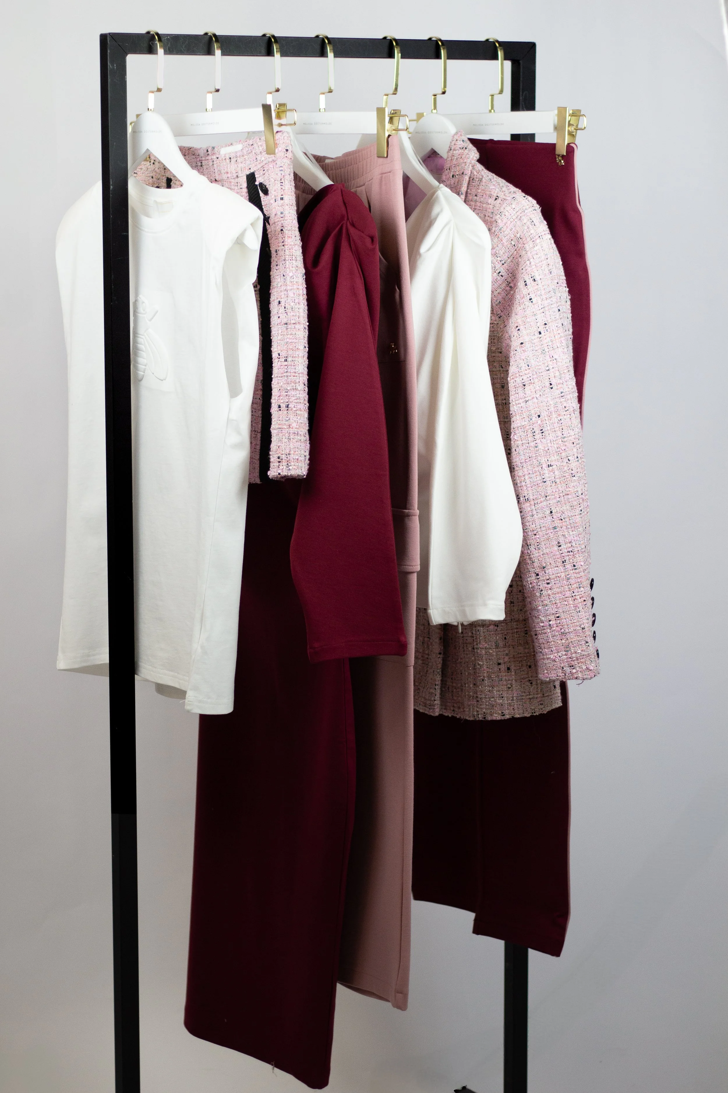 A clothing rack with white, pink, and burgundy garments, including skirts, pants, and tops, arranged on white and gold hangers.