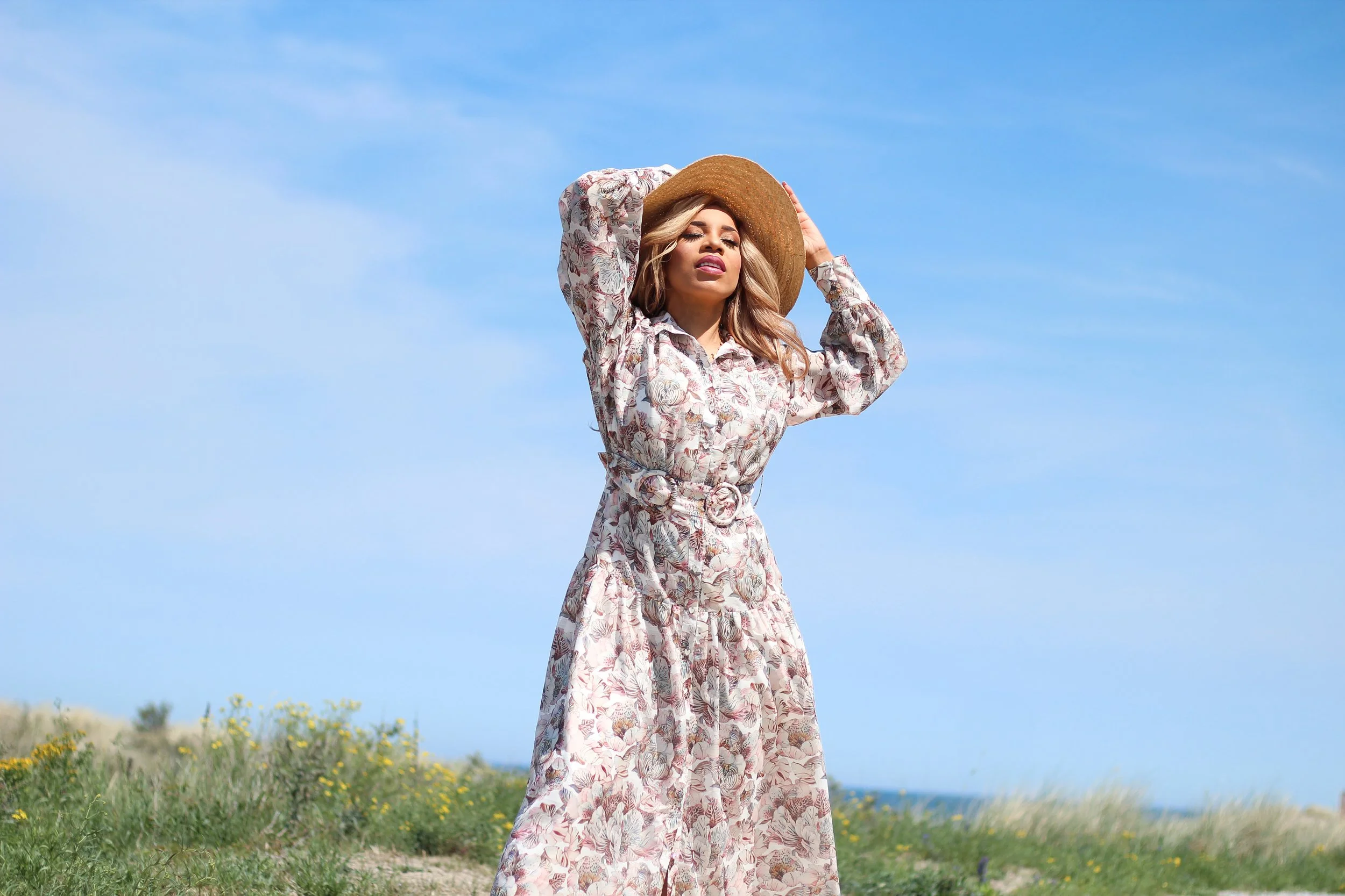 Woman in a floral dress and wide-brimmed hat standing outdoors on a sunny day, with a blue sky and grassy landscape in the background.