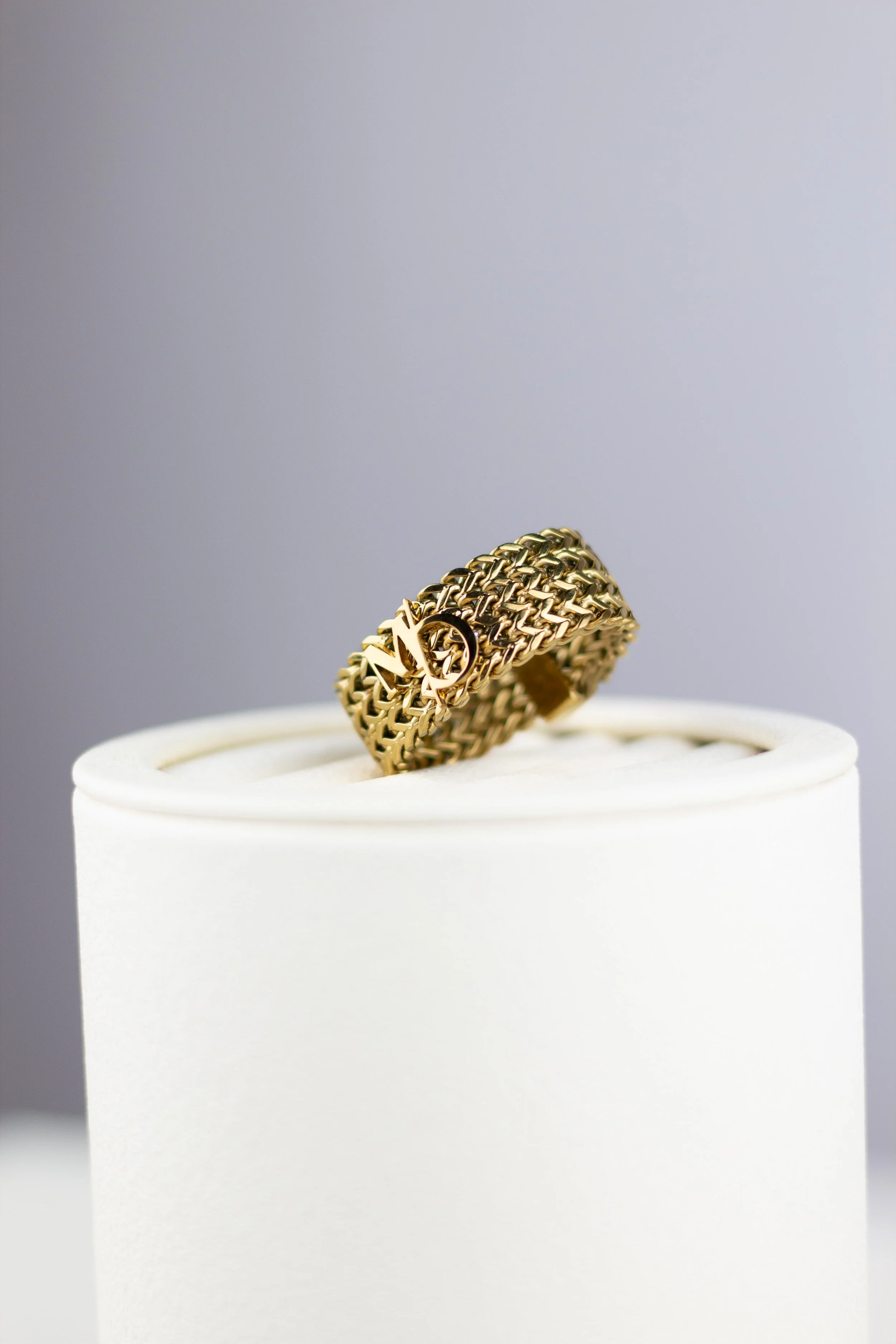 A gold ring with a textured woven design and a prominent logo, displayed on a white ring holder.