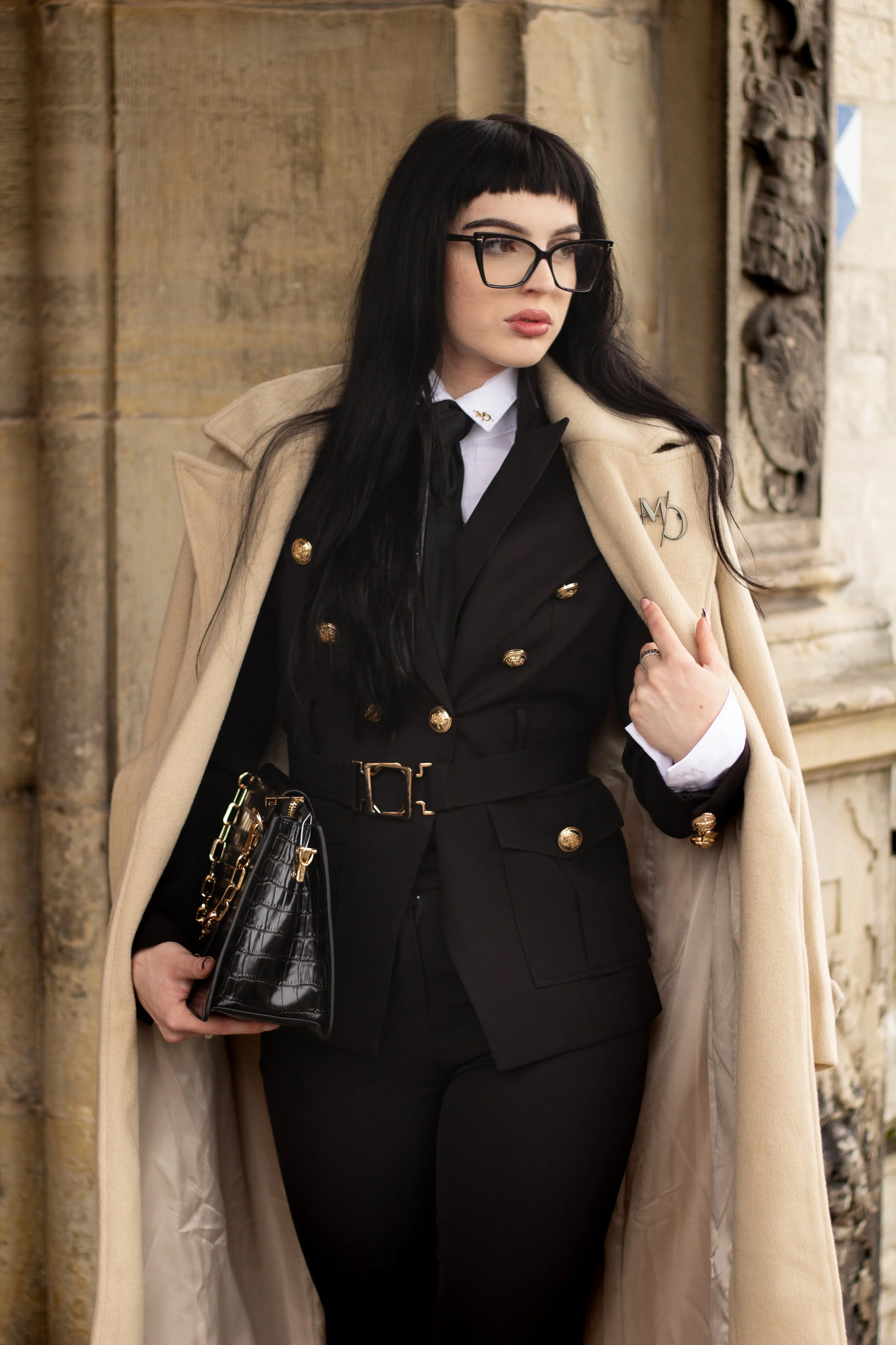 A woman with long dark hair, wearing glasses and a stylish coat, standing against a stone wall, dressed in a black double-breasted suit with gold buttons, holding a black handbag with gold chain accents.