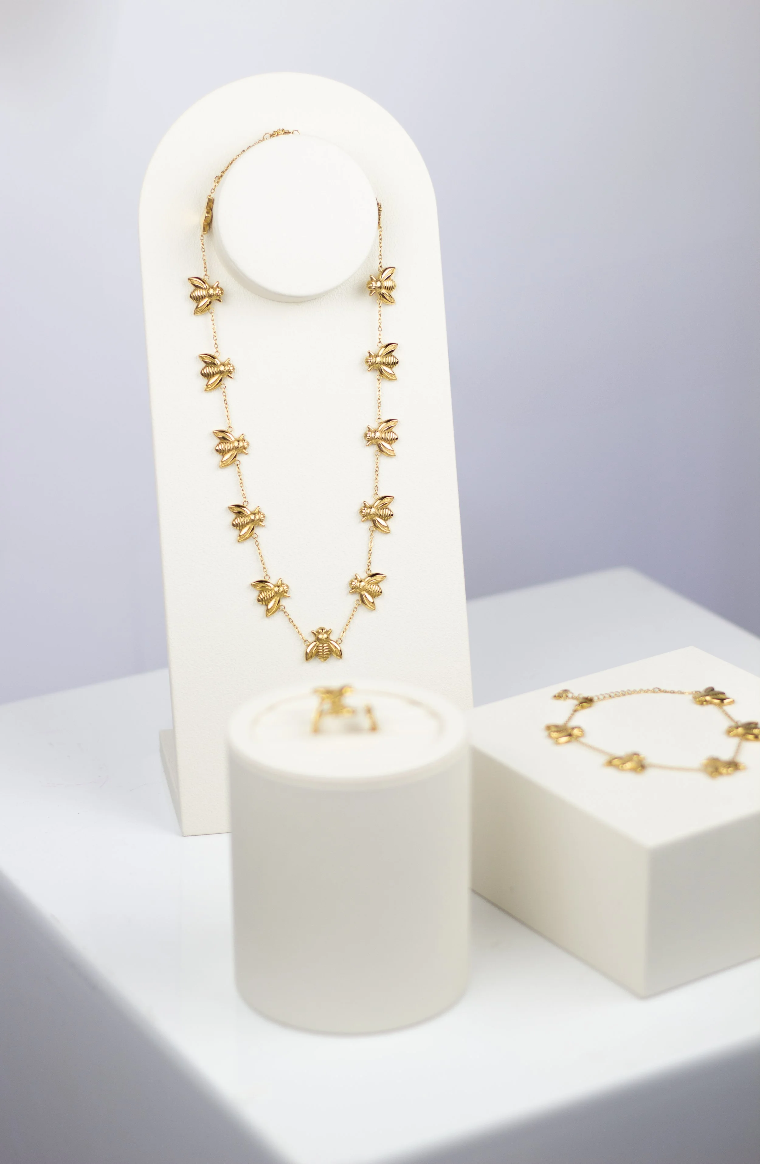 Gold jewelry display featuring a necklace with bee charms, a matching bracelet, and another piece with bee charms, all set on white stands and platforms.
