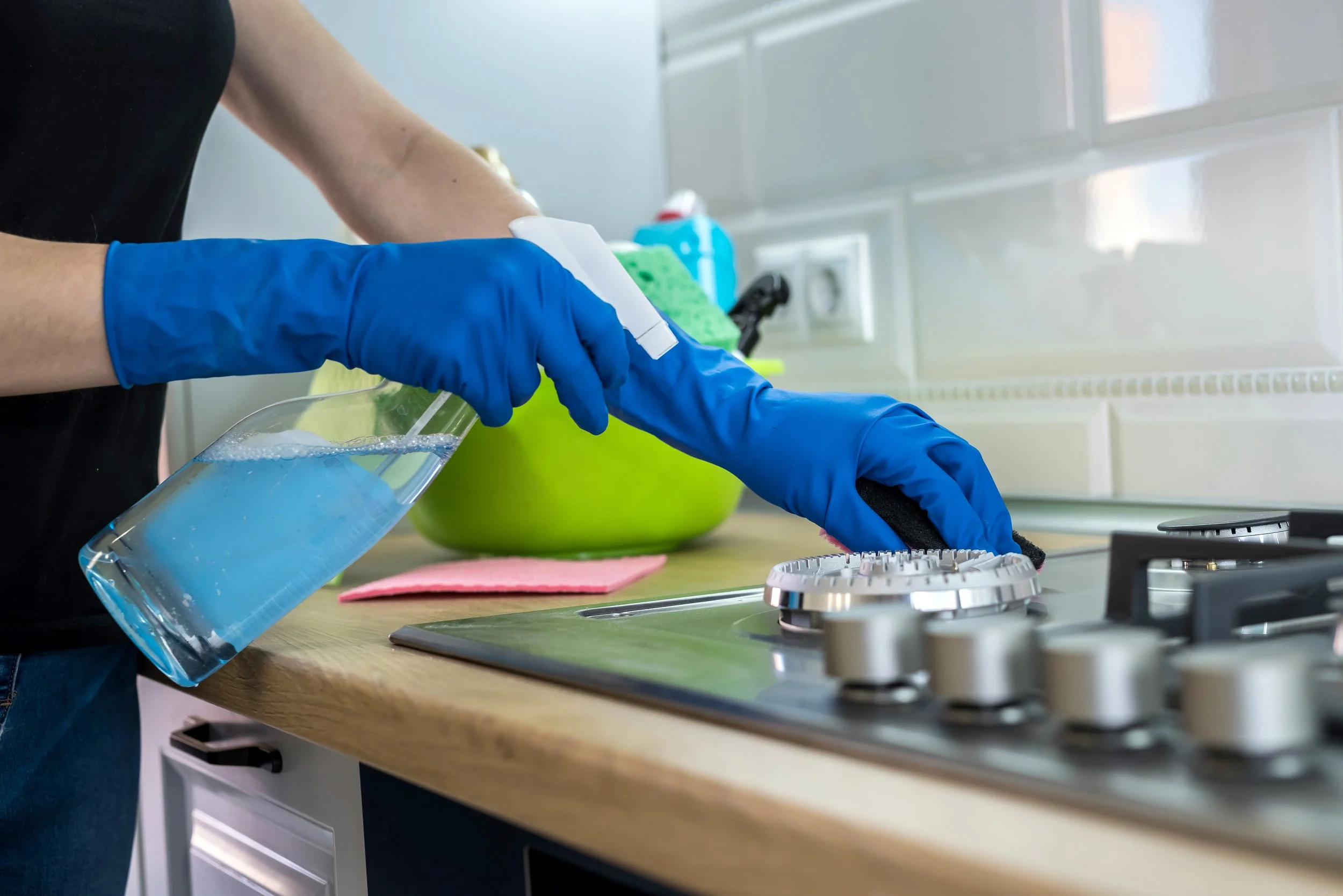 A person wearing blue gloves cleaning a stovetop with a spray bottle and cloth in a kitchen.