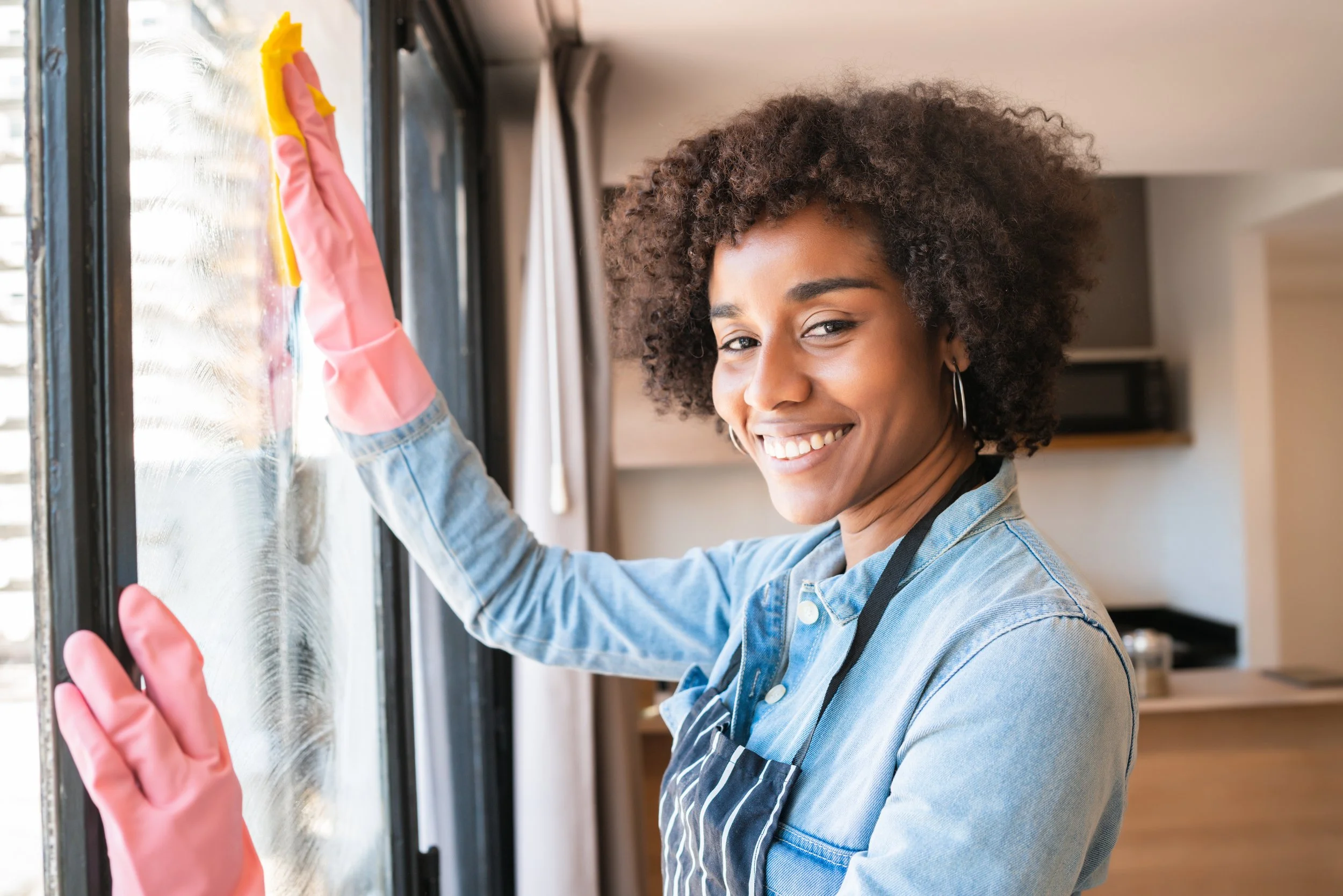 A woman wearing pink rubber gloves and an apron cleaning a window with a yellow sponge in her hand.