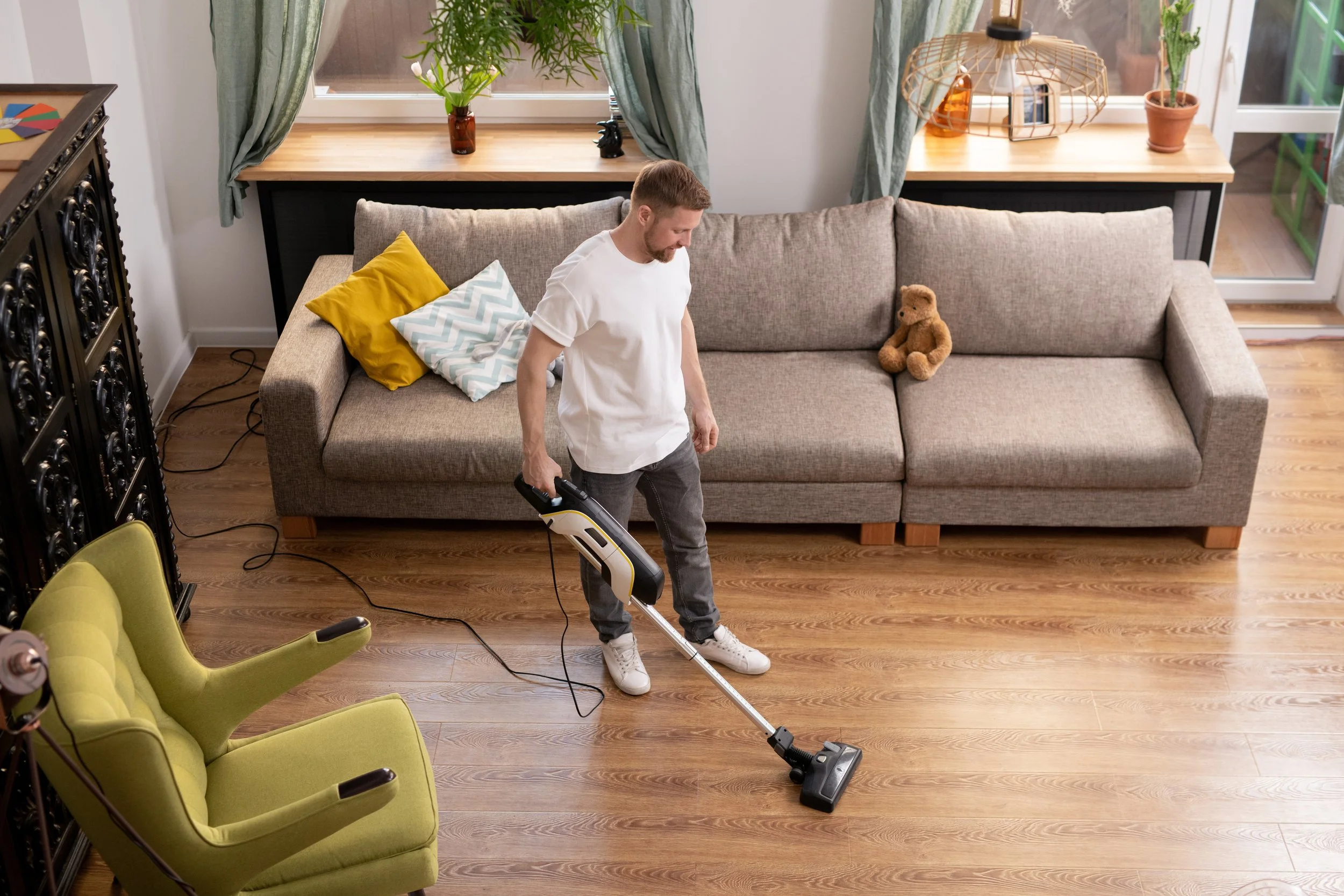 A man using a cordless vacuum cleaner to clean a wooden floor in a living room with a gray couch, a teddy bear, throw pillows, and a window with curtains.